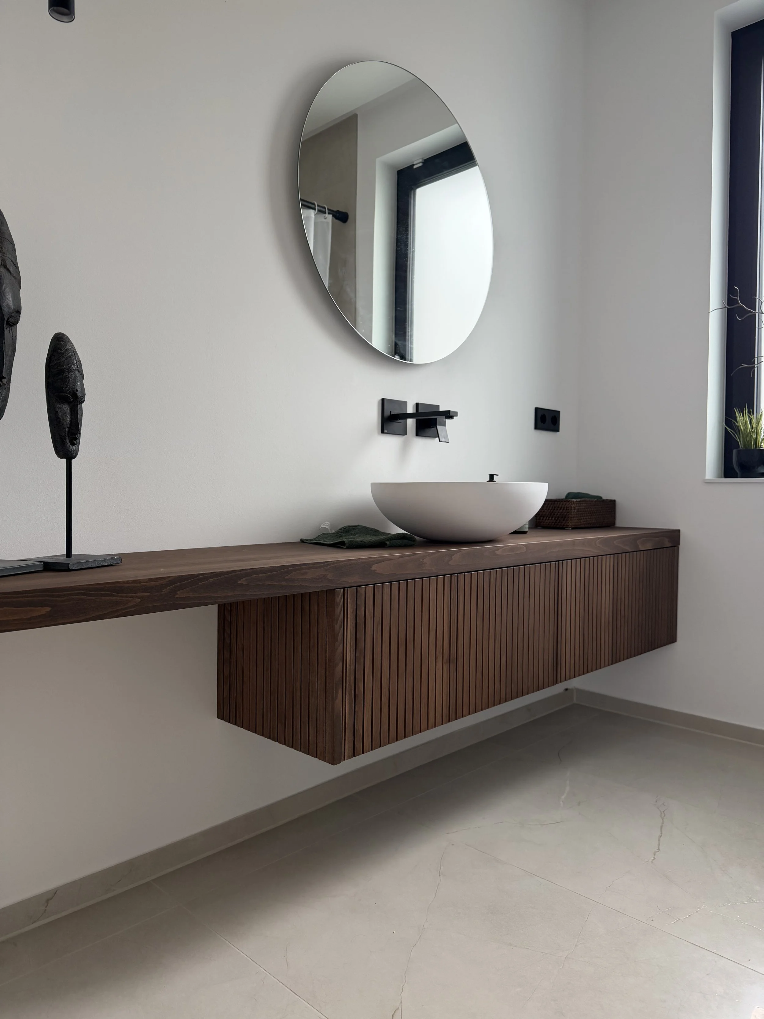 Moderner Badezimmerwaschtisch mit weißer runder Schüssel, schwarzer Wasserhahn, Waschbecken und einem großen ovalen Spiegel an einer weißen Wand. Es gibt Kunstfiguren aus Holz auf der linken Seite auf einer Holzablage. Ein Bürofenster an der rechten Seite lässt natürliches Licht hereinströmen.