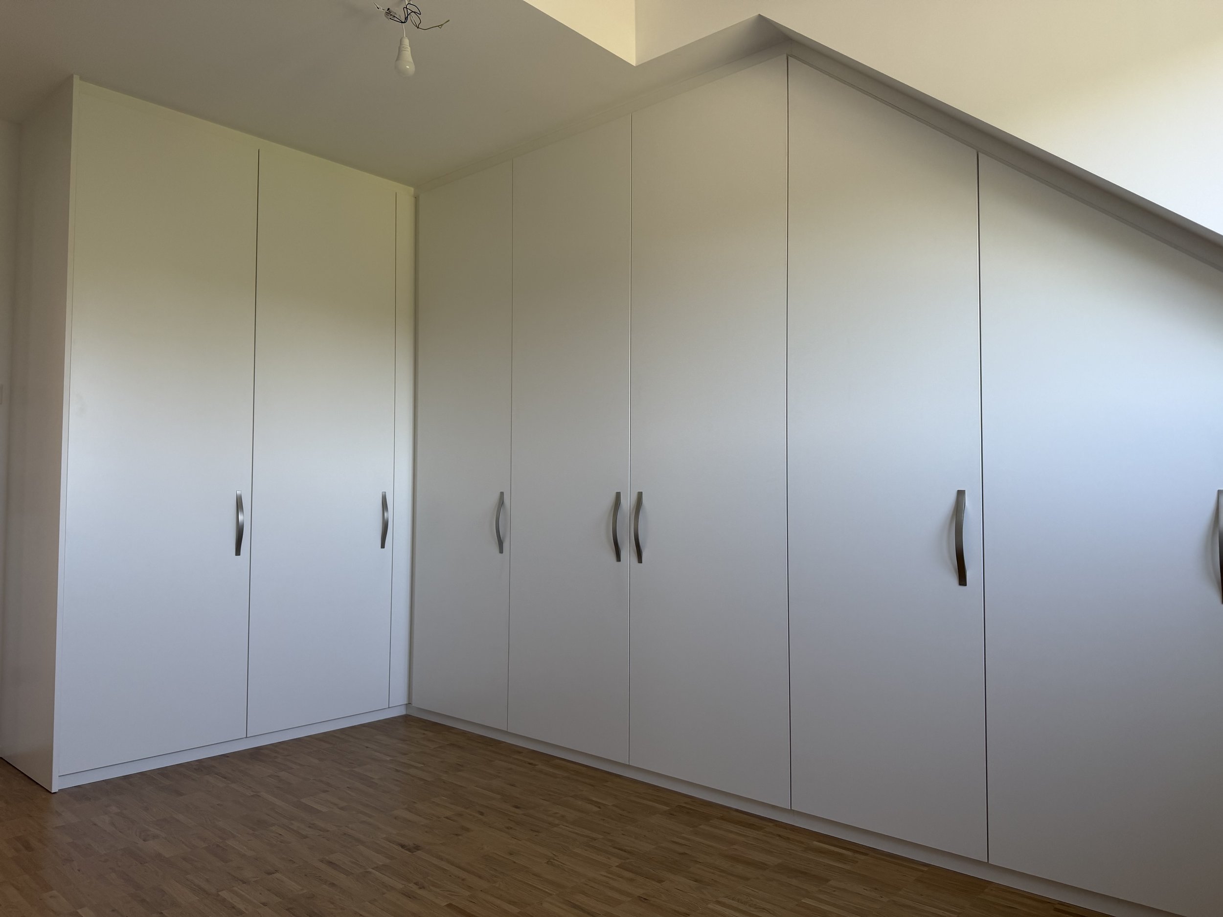 Weiße Einbauschränke mit geschwungenen Griffen in einem Raum mit Holzboden, an einer Wand mit Schrägdach.