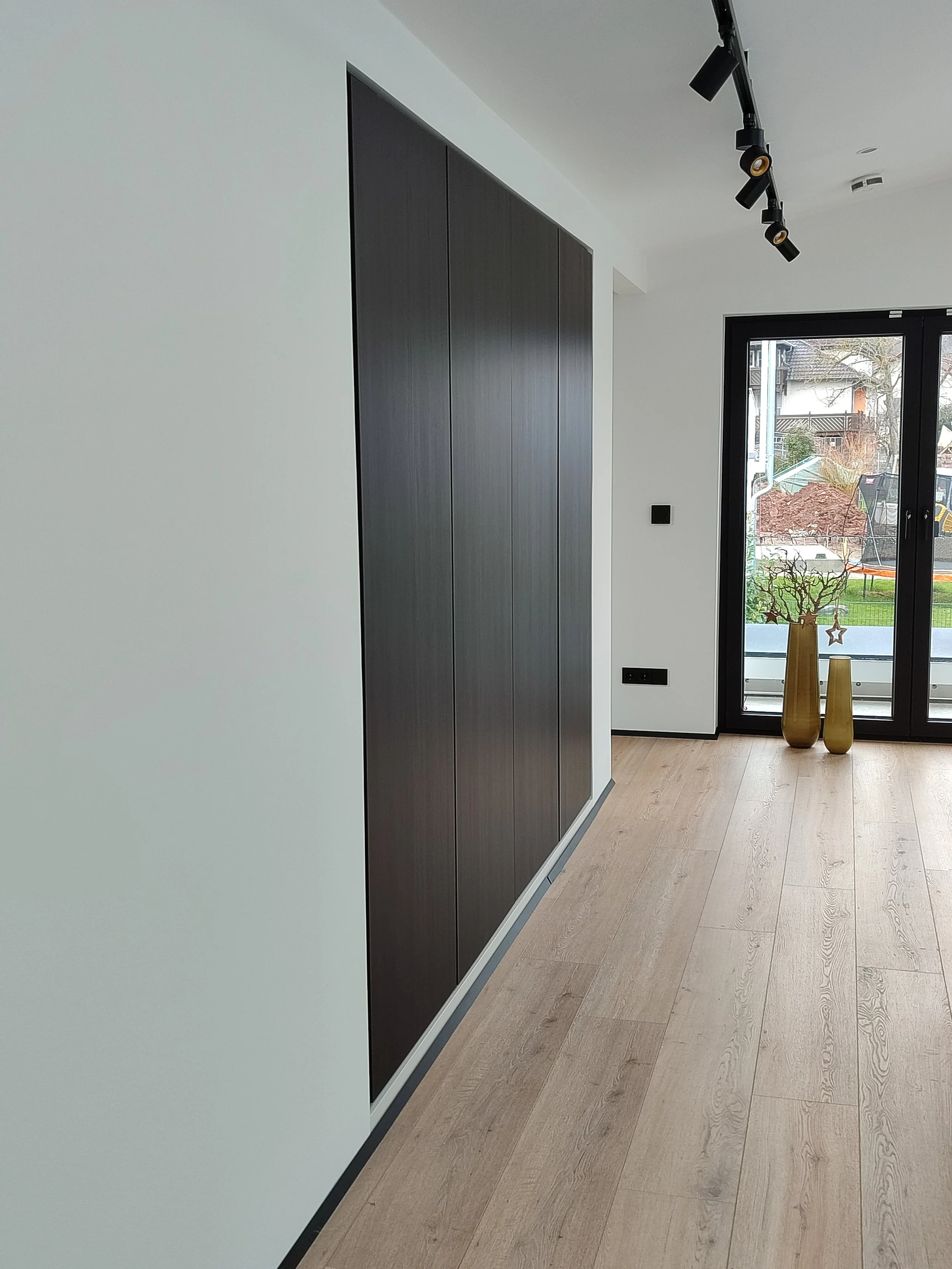 Moderner Raum mit hellen Wänden, schwarzer Schiebetür, Holzboden, großer Fensterscheibe mit Blick auf einen Garten.