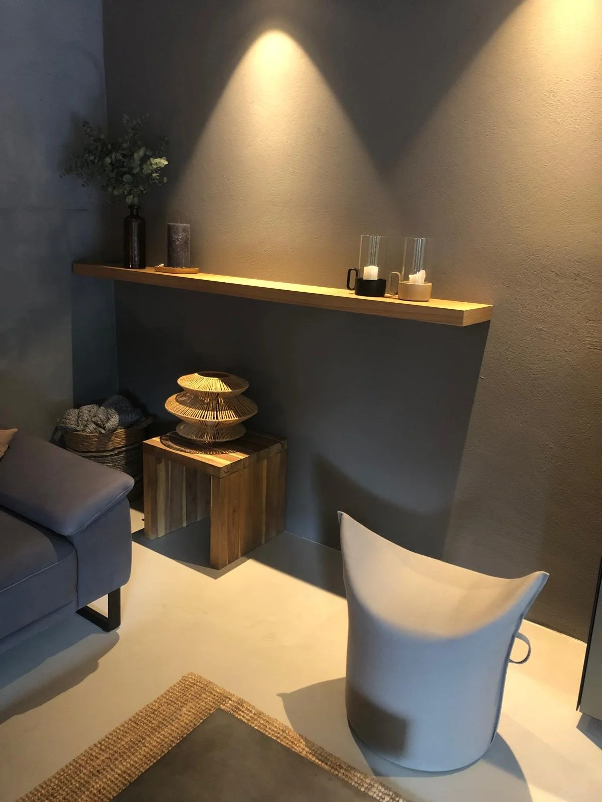 Ein Wohnzimmer mit einer grauen Couch, einem weißen Stuhl, einer Holzseite aus, einer Wandlampe, einer Wanddekoration mit Kerzen, einer Vase mit Pflanzen auf einem Regal, und einem dekorativen Holzobjekt auf einem Holztisch.