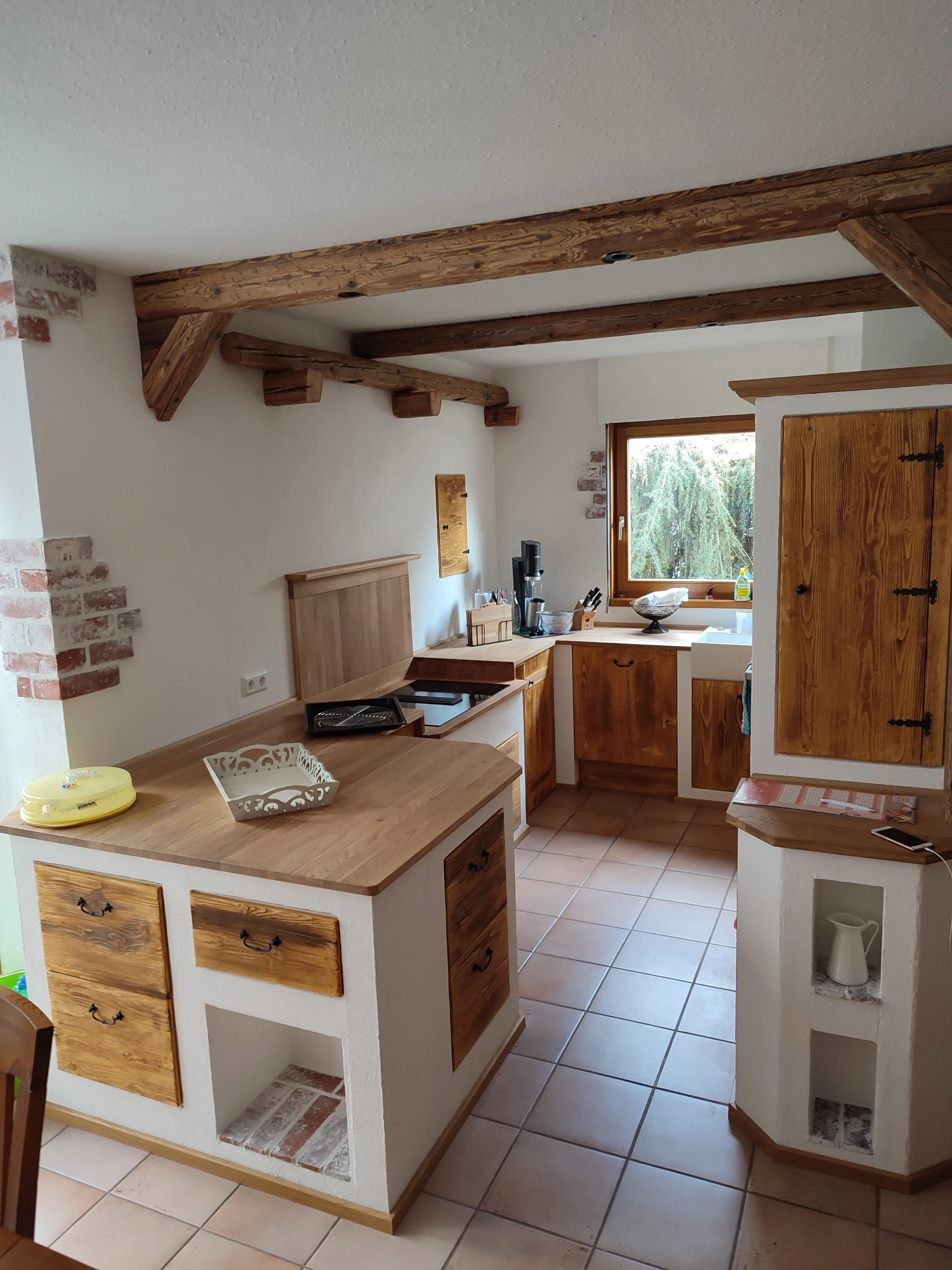 Eine Küchenecke mit Holzmöbeln, einer Arbeitsplatte, einem Fenster, einer kleinen Theke und einigen Gegenständen auf der Arbeitsfläche, mit Blick auf Fliesenboden und Holzbalken an der Decke.