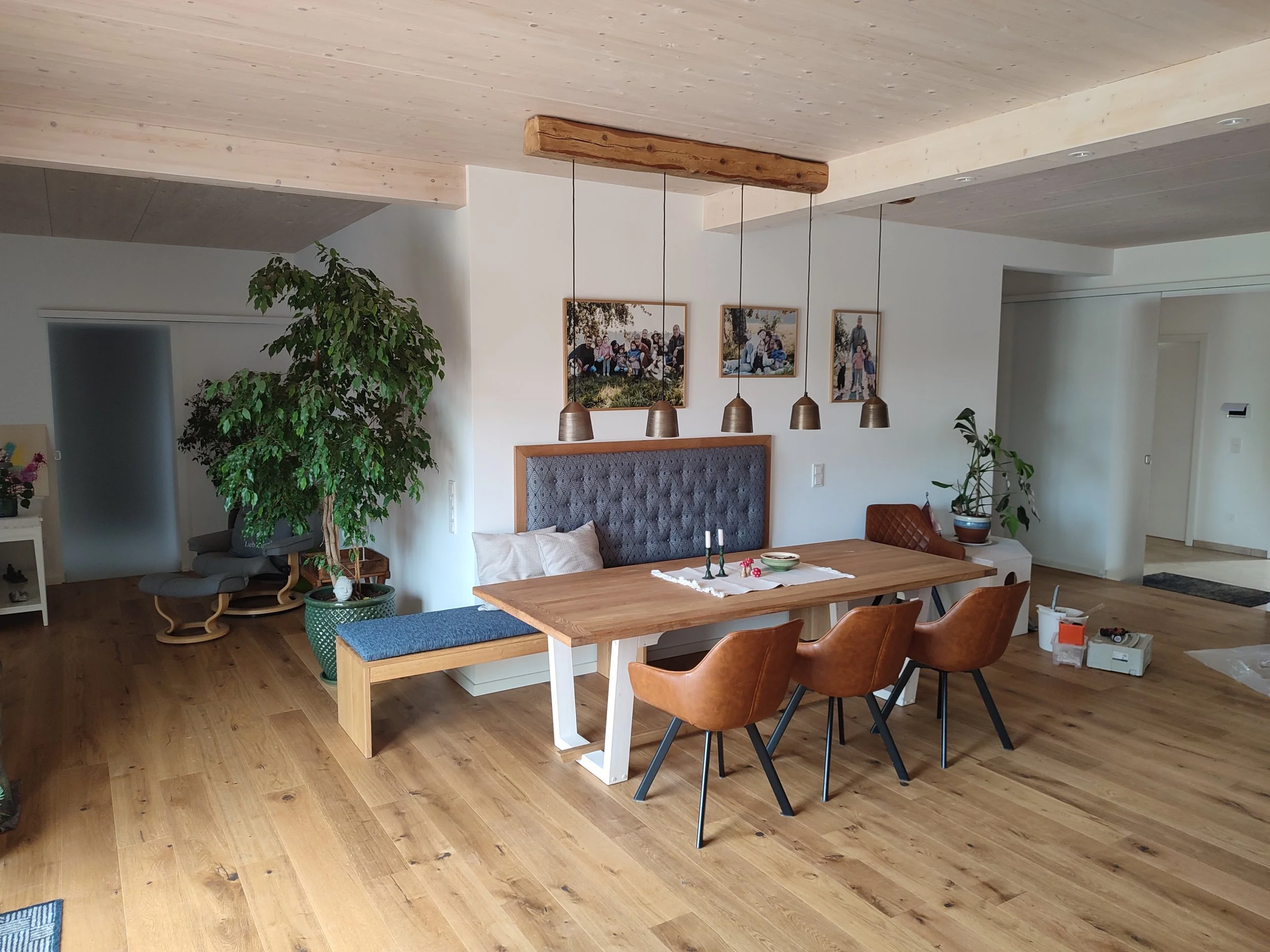Innenansicht eines modernen Wohnzimmers mit Holzdecke, einer großen grünen Pflanze, Wandbildern, Esstisch mit mehreren Holzstühlen und einer Wand mit integriertem Kamin.