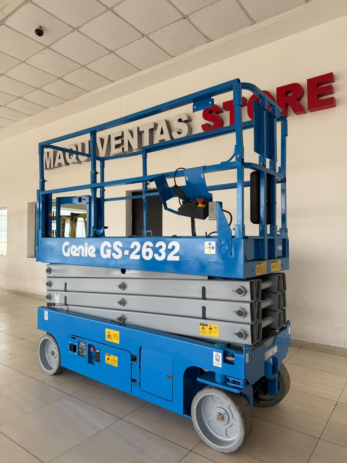 Plataforma elevadora azul Marca Genie modelo GS-2632 en interior de edificio con paredes blancas y letrero en la pared que dice "MAQUINAS VENTAS STORE" en letras negras y rojas.