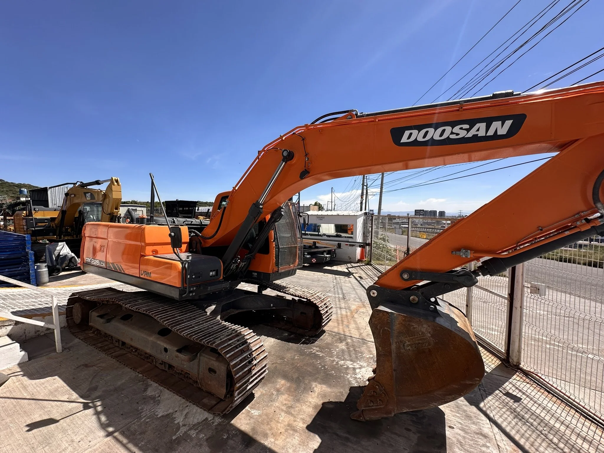 Excavadora de color naranja con la marca Doosan, ubicada en un sitio de construcción al aire libre bajo un cielo despejado.