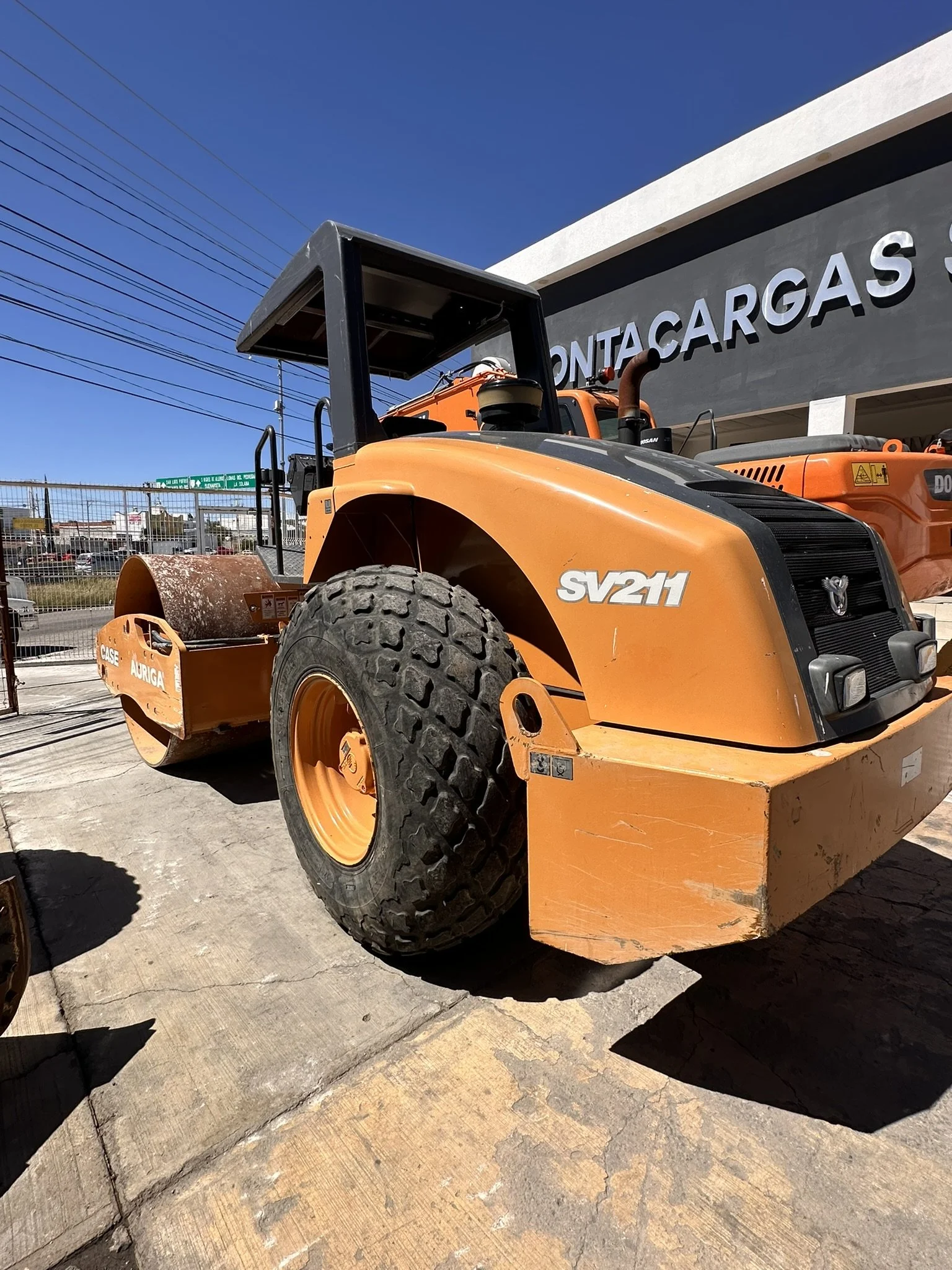 Maquinaria de construcción, rodillo compactador, de color naranja, estacionado en un lote con un fondo de edificio y cielo despejado.
