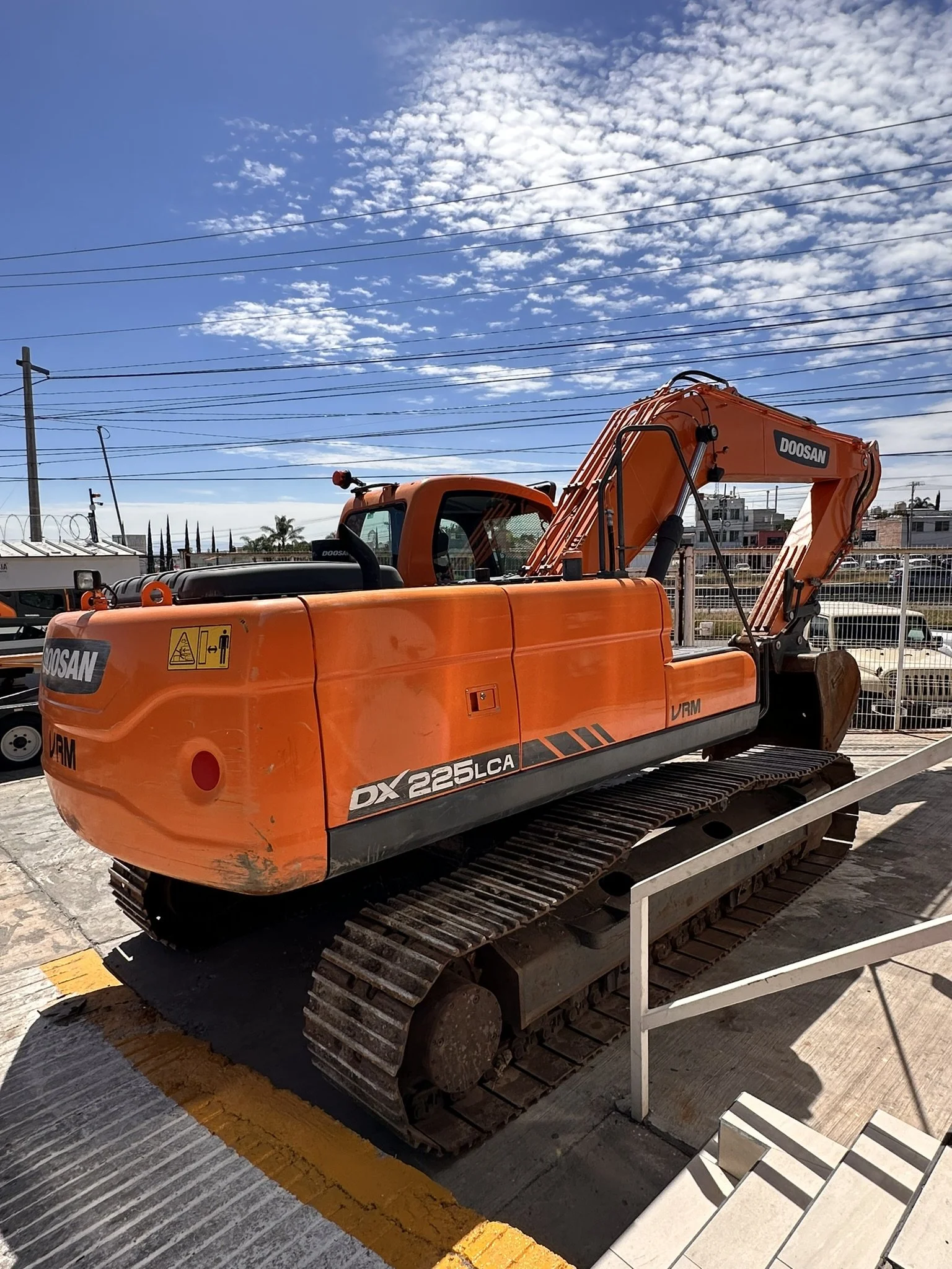 Excavadora naranja de la marca Doosan en exhibición en un área al aire libre, con una cerca de fondo y cielo despejado con nubes pequeñas.