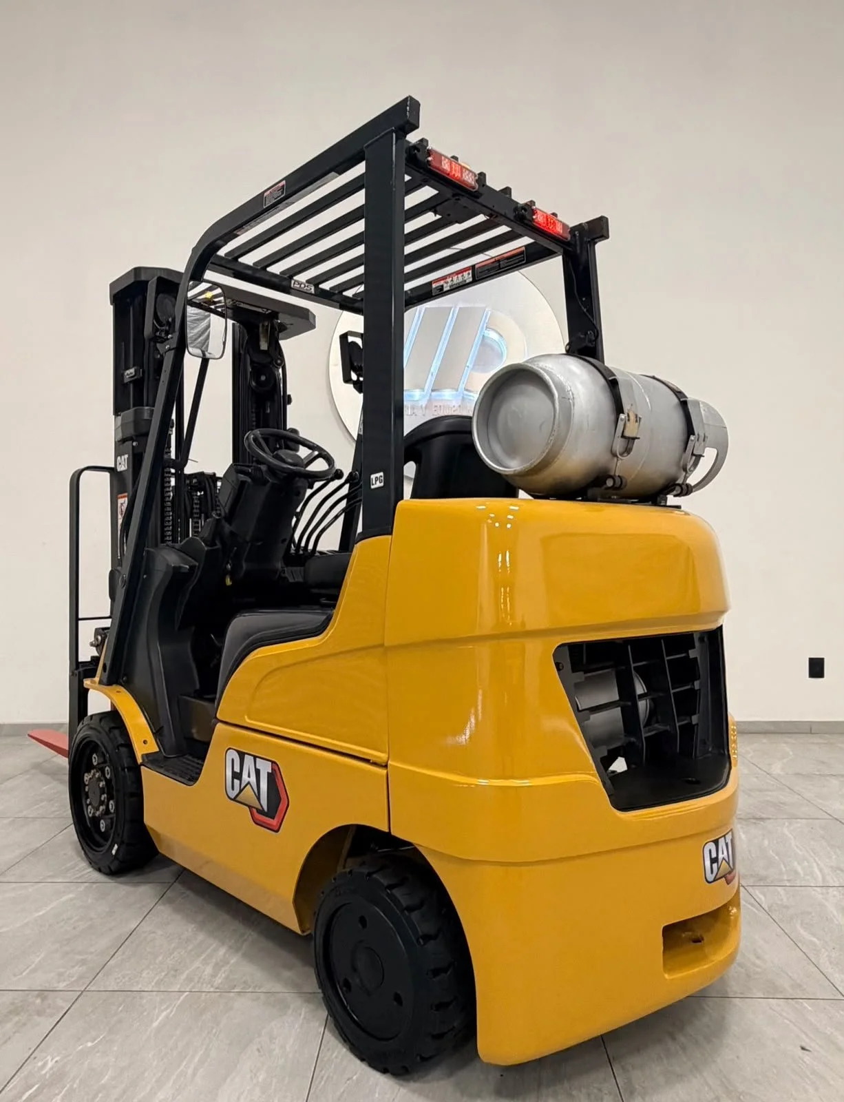 Montacargas amarillo de la marca Caterpillar con cilindro de gas en la parte trasera, en interior de edificio con pared blanca y suelo de baldosas.