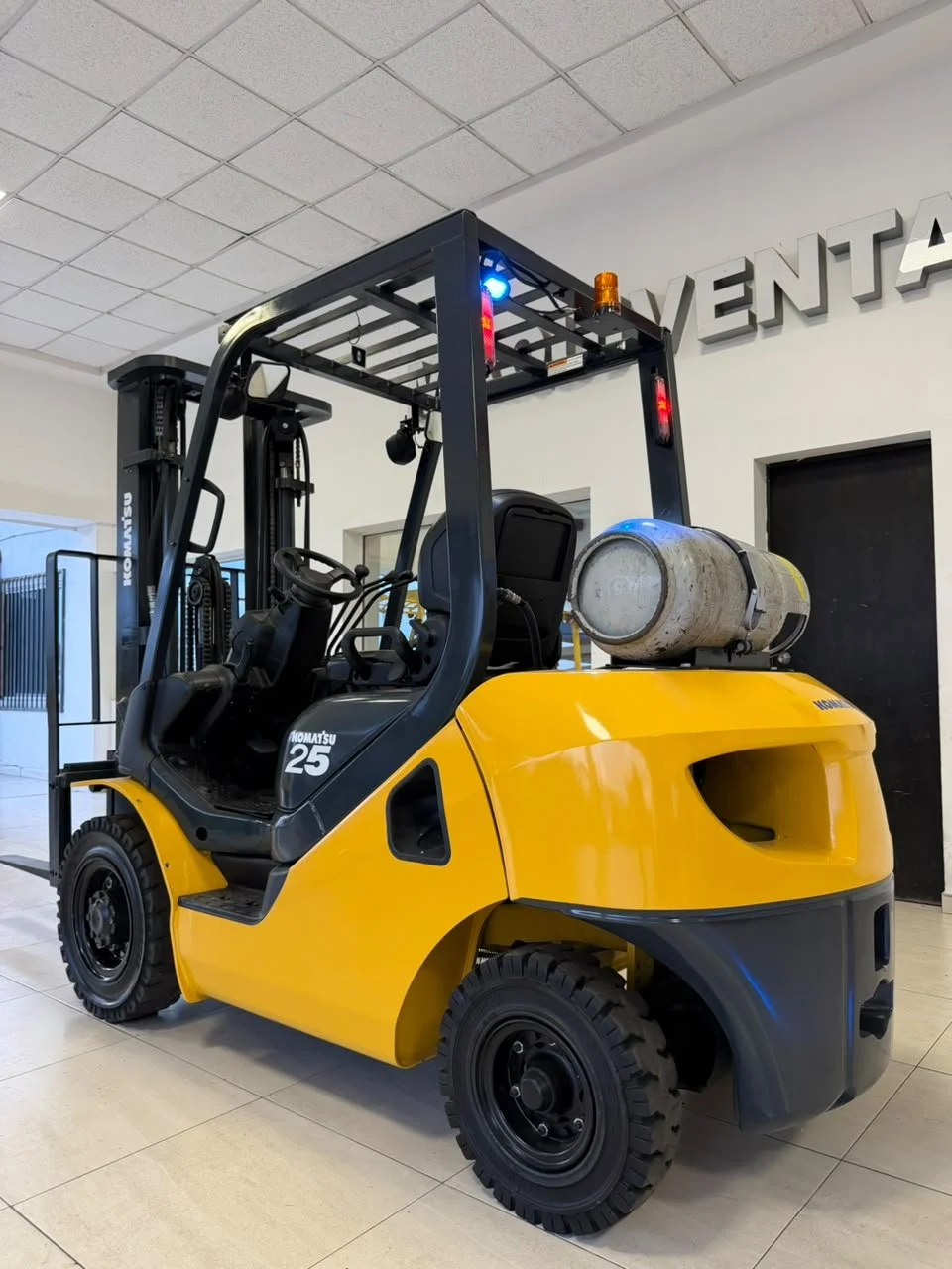 Montacargas amarillo con cilindro de gas en la parte trasera, ubicado en interior de edificio con techo de paneles blancos, piso de baldosas y paredes blancas con letras que dicen 'VENTA'.