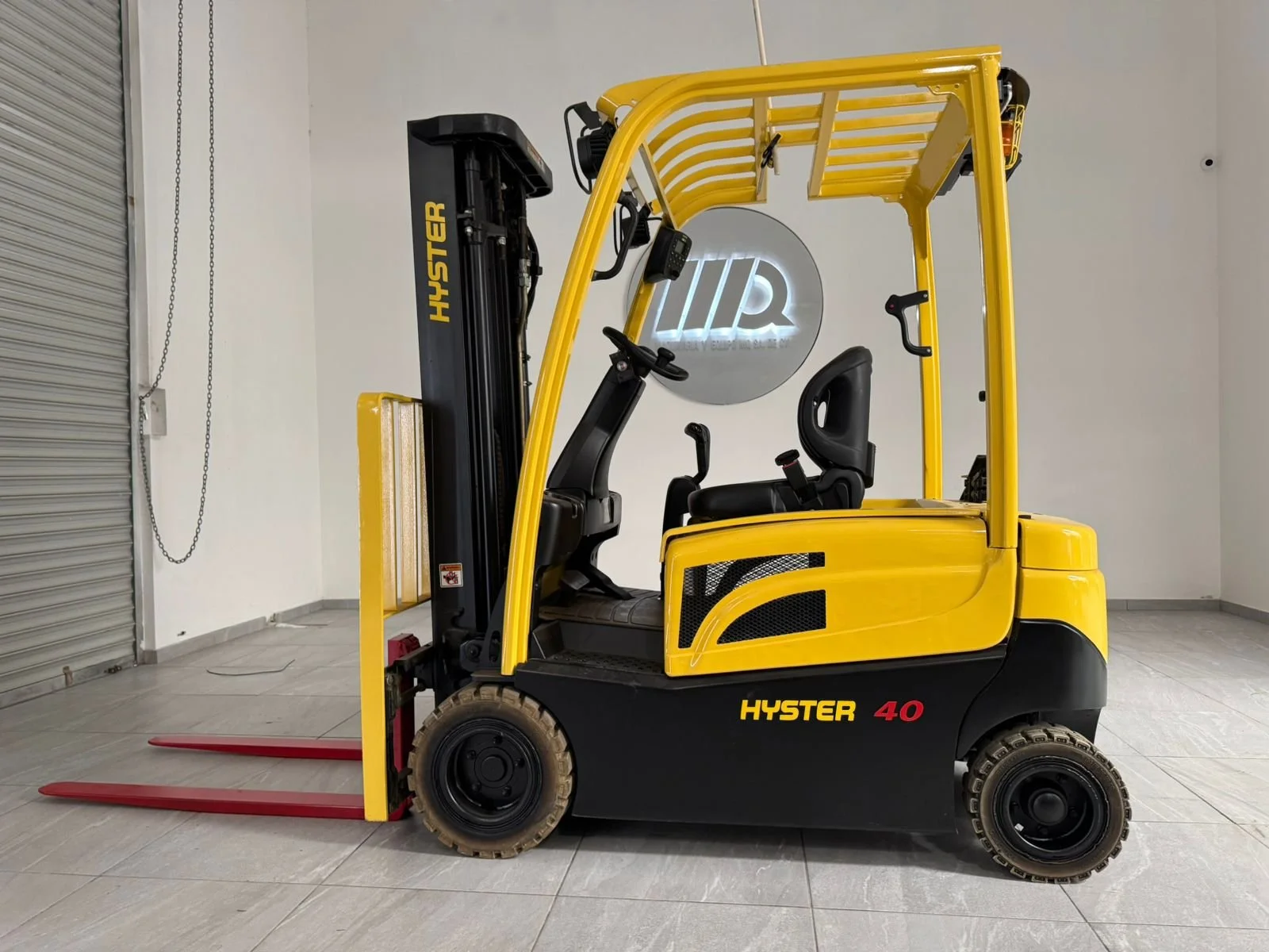 Montacargas amarillo y negro en interiores con paredes blancas y suelo de baldosas, logotipo iluminado de HQR en la pared.