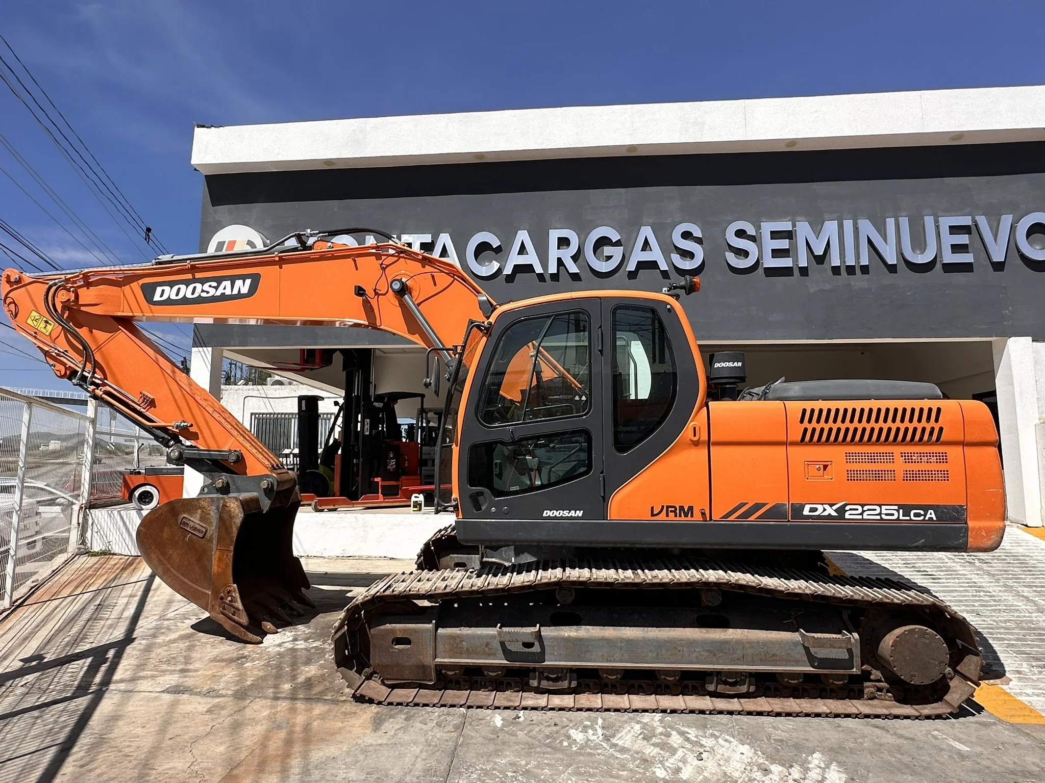 Excavadora orange de la marca Doosan en un terreno con una construcción de fondo que tiene un letrero que dice 'CARGAS SEMINUEVOS'. La excavadora está sobre una pendiente de concreto y tiene una cuchara grande en el extremo del brazo.