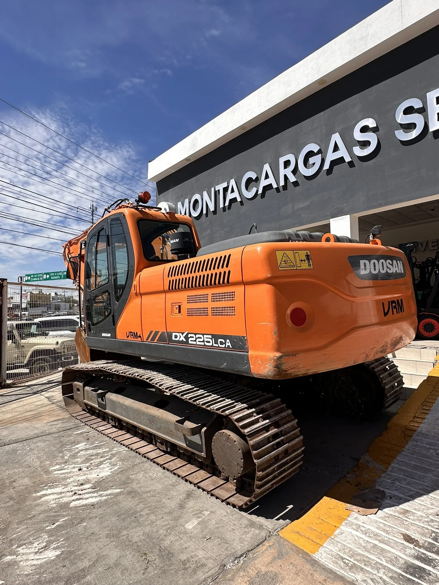 Excavadora marca Doosan de color naranja en exhibición frente a un edificio con cartel que dice Montacargas.