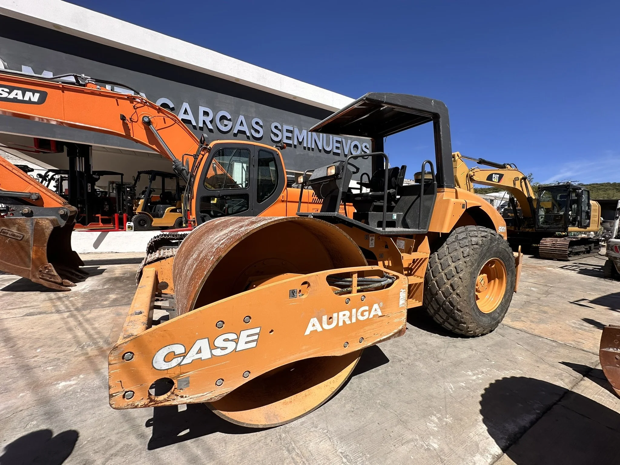 Rodillo compactador en un terreno de trabajo, con maquinaria pesada en el fondo y un edificio de ventas de maquinaria en el fondo.