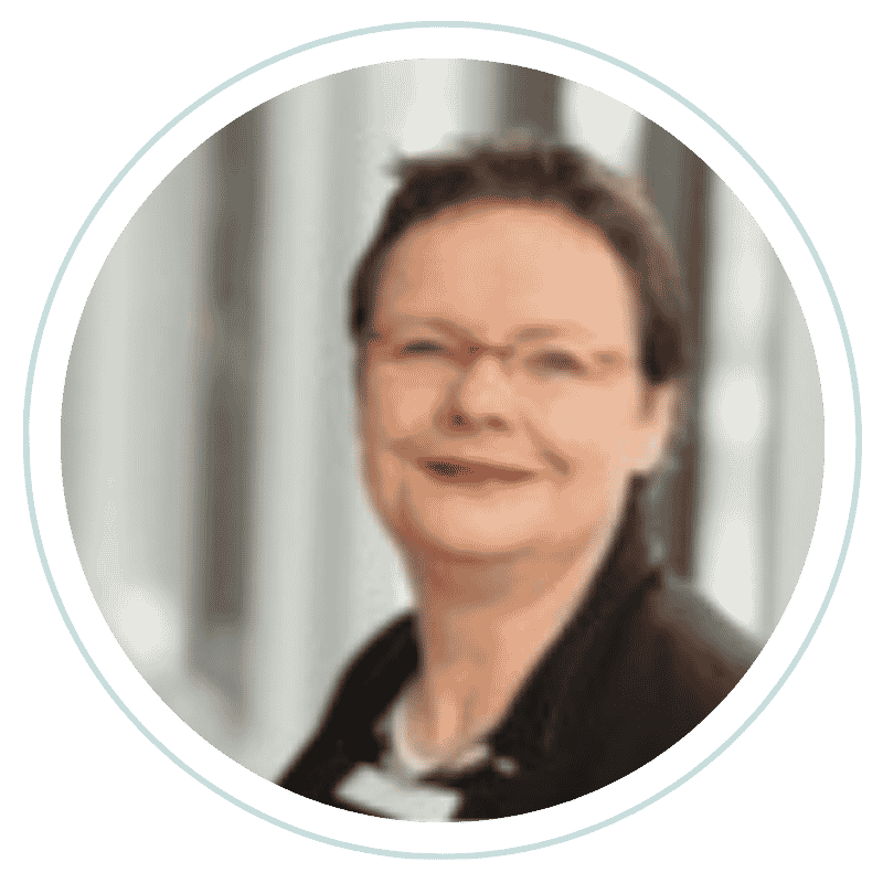 Ein Foto einer lachenden Frau mit Brille, dunklen Haaren und dunkler Jacke, aufgenommen in einer indoor-Umgebung.