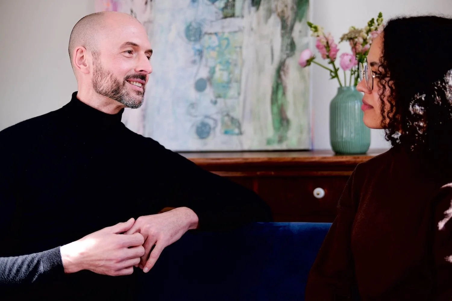 Ein Mann und eine Frau sitzen einander gegenübers, halten die Hände und sprechen miteinander, im Hintergrund ein Wandbild und eine Vase mit Blumen.