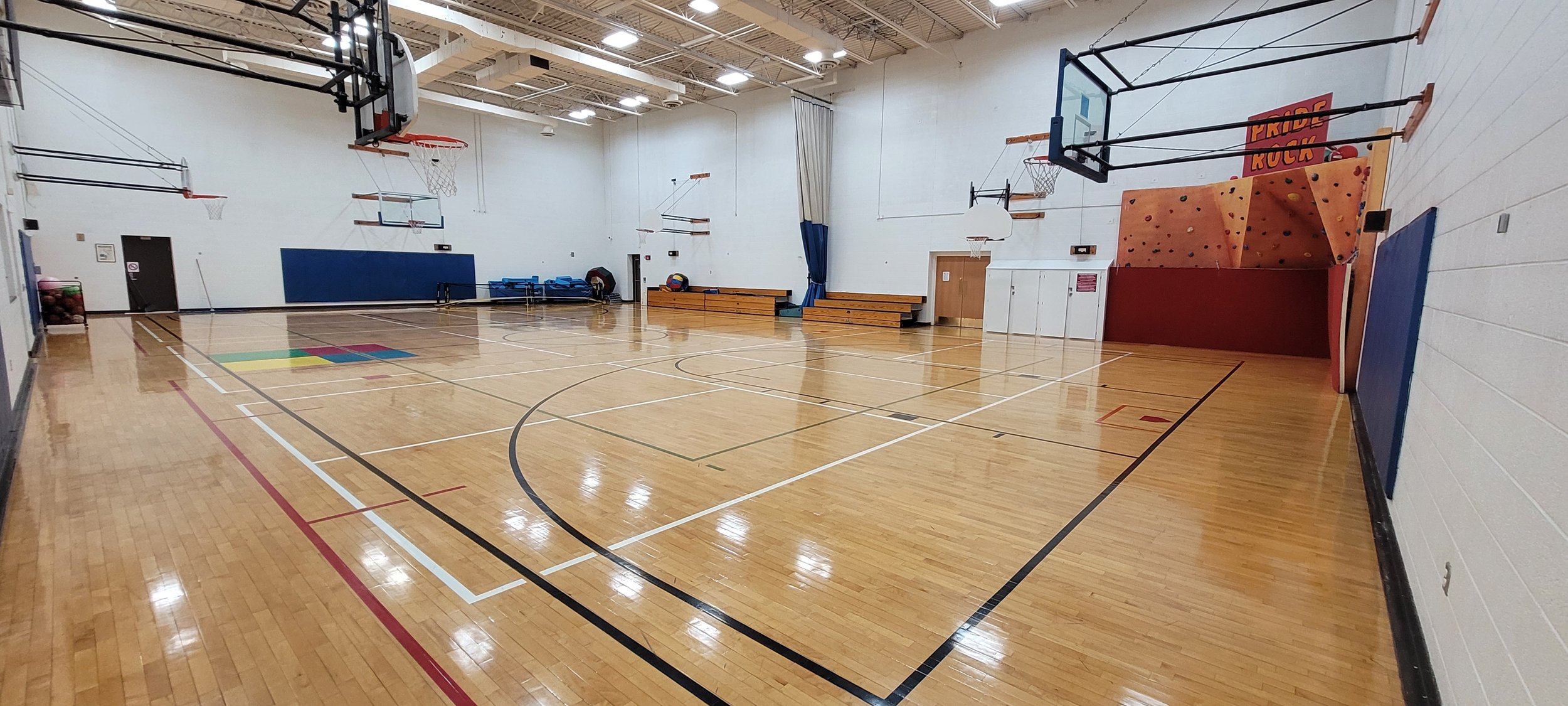 A gymnasium with short climbing wall, basketball hoops, and short bleachers on one side.