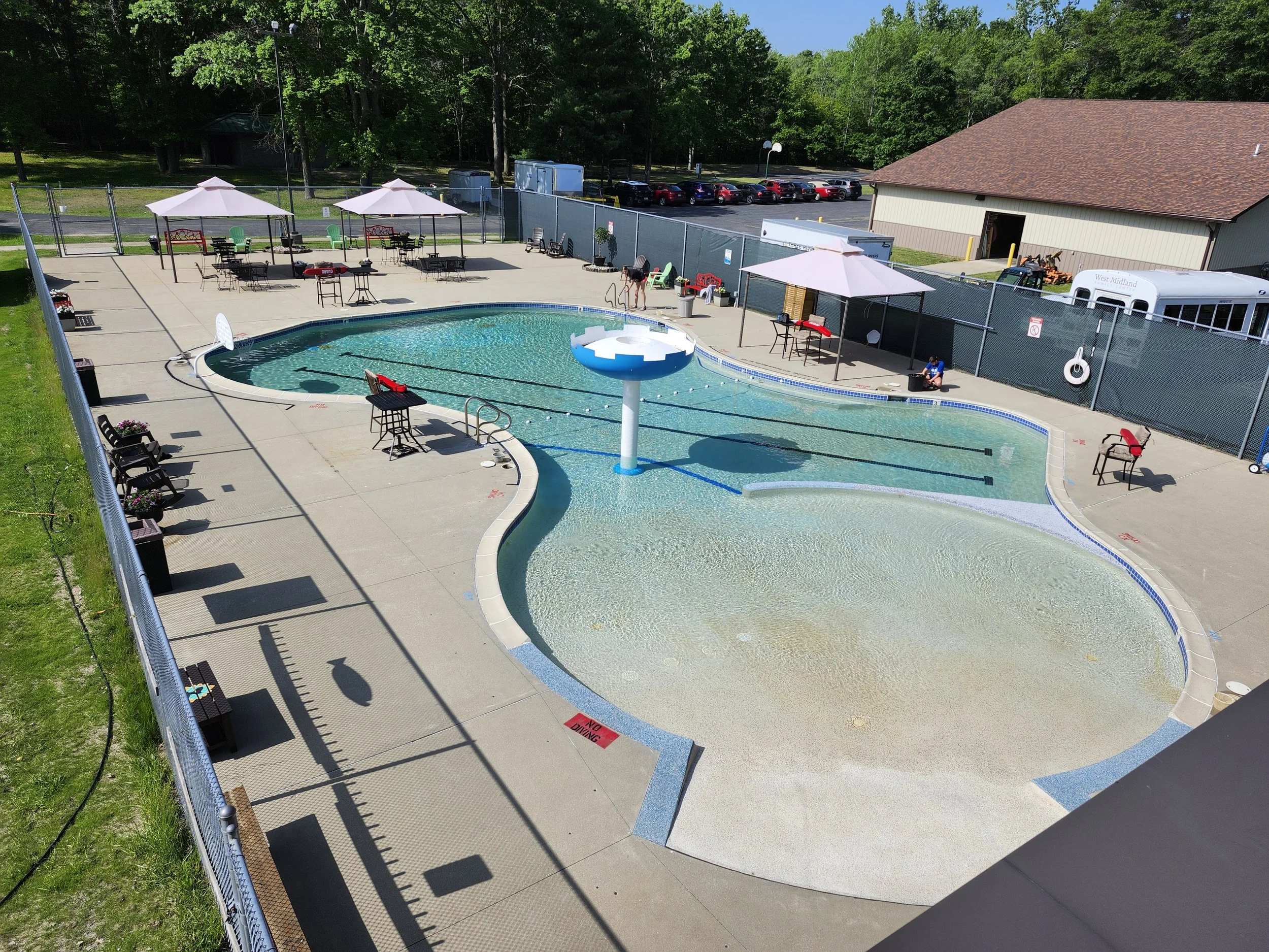 A large outdoor pool in a fenced-in area, with canopies, benches, tables, and chairs on the cement pool deck