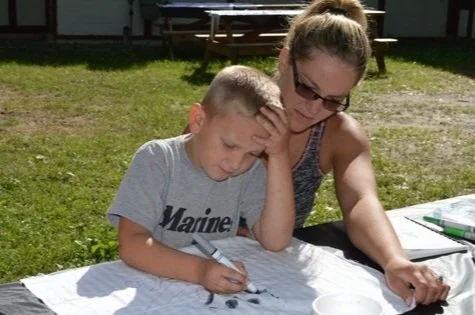 A young boy colors a page with a marker as his mother looks over his shoulder. They are sitting at a picnic table outdoors