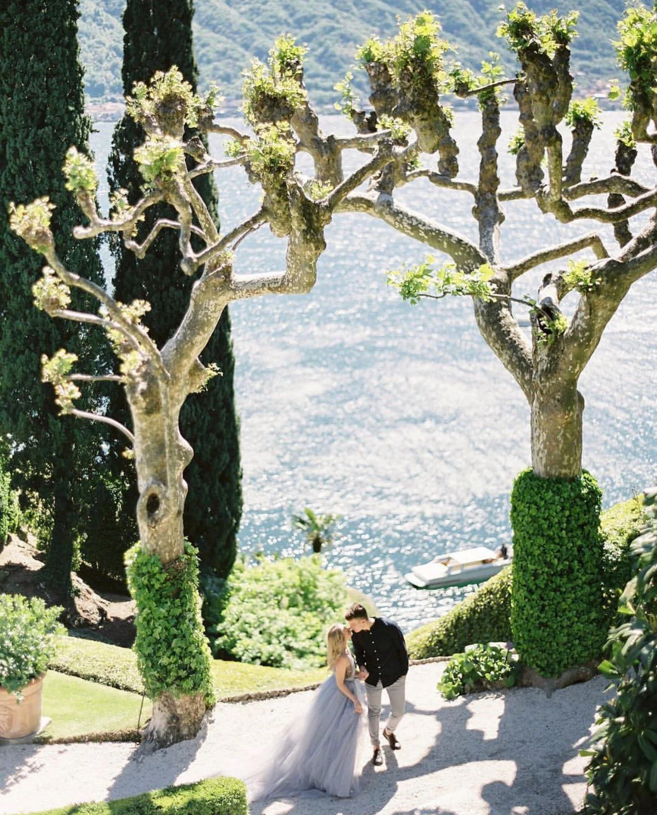A couple dancing outdoors near a lake, surrounded by trimmed trees and lush greenery, with a boat floating on the water in the background.