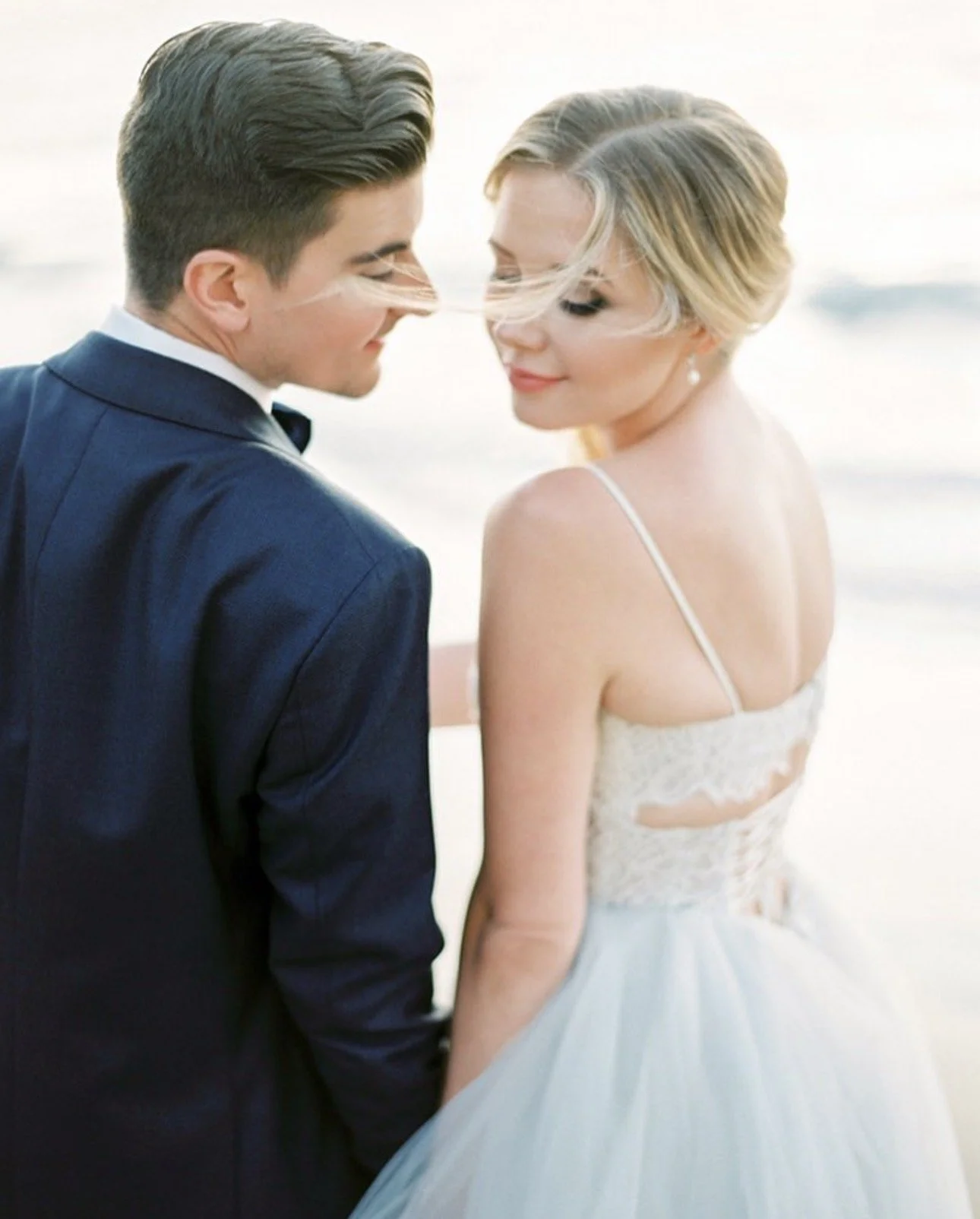 A happy bride and groom stand close together on the beach during sunset, gazing affectionately at each other.