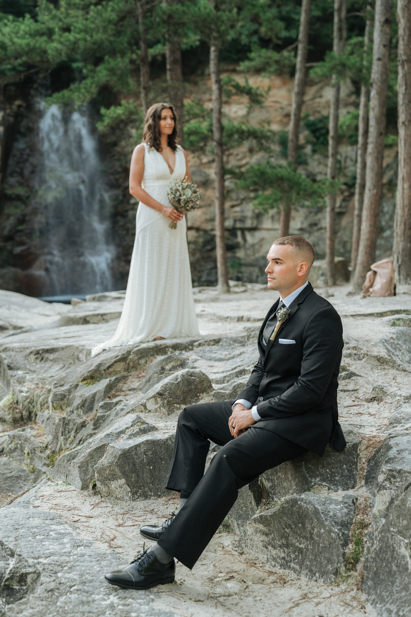 A bride standing in a white dress holding a bouquet, looking at a groom sitting on a large rock, dressed in a black suit and tie, outdoors near a waterfall and surrounded by trees.