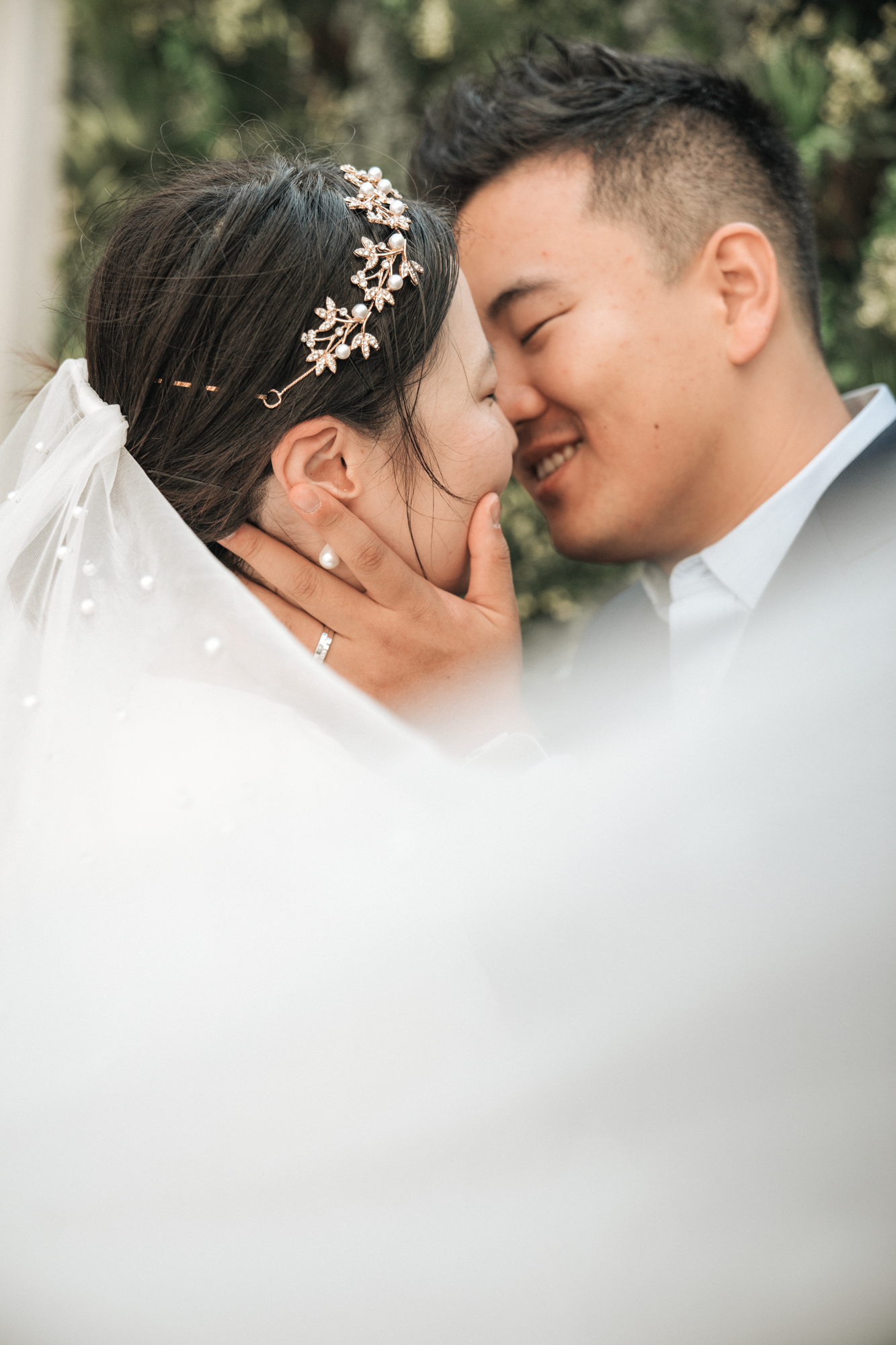 A bride and groom sharing a close, intimate moment, touching foreheads and holding each other's faces, outdoors in a natural setting.