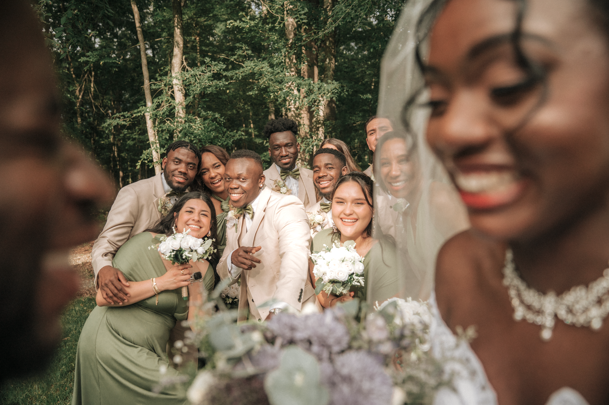 A group of wedding guests posing for a photo outdoors in a forest, with the bride on the right side smiling, and the rest of the guests, including a woman holding a bouquet of white flowers, gathered together behind her.