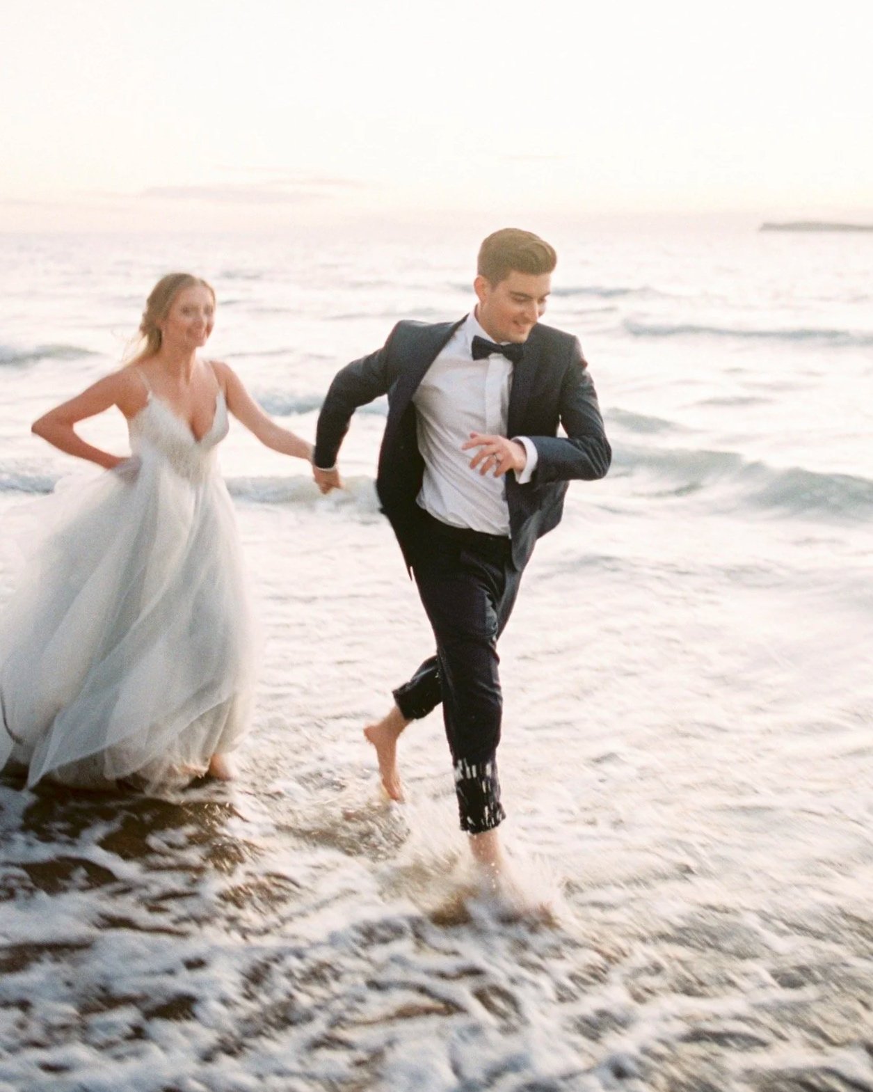 A bride and groom running barefoot along the beach at sunset, dressed in wedding attire with the bride in a white gown and the groom in a black tuxedo.