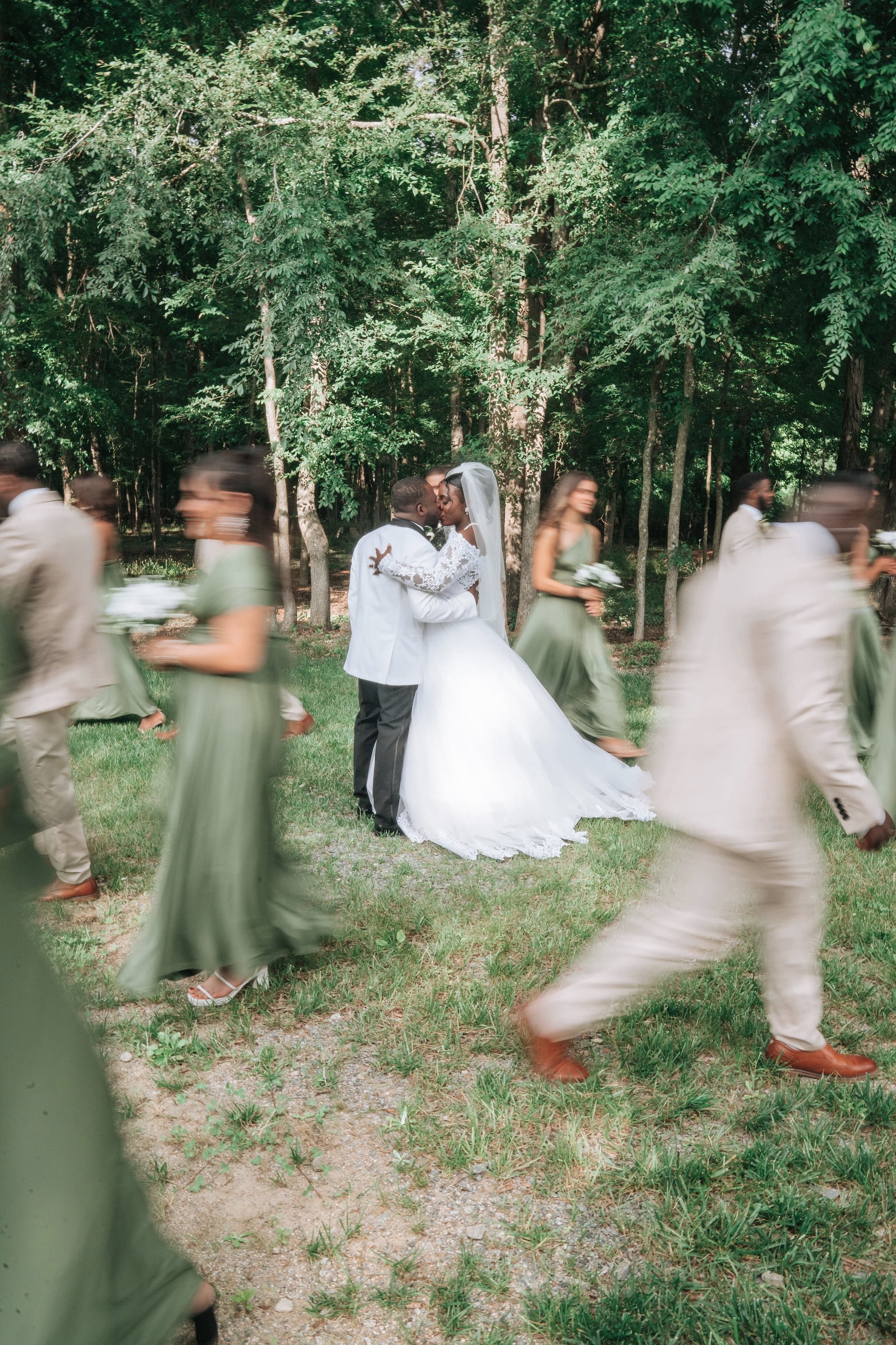 Wedding celebration outdoors with a bride and groom kissing, surrounded by bridesmaids and groomsmen walking in a forested area.
