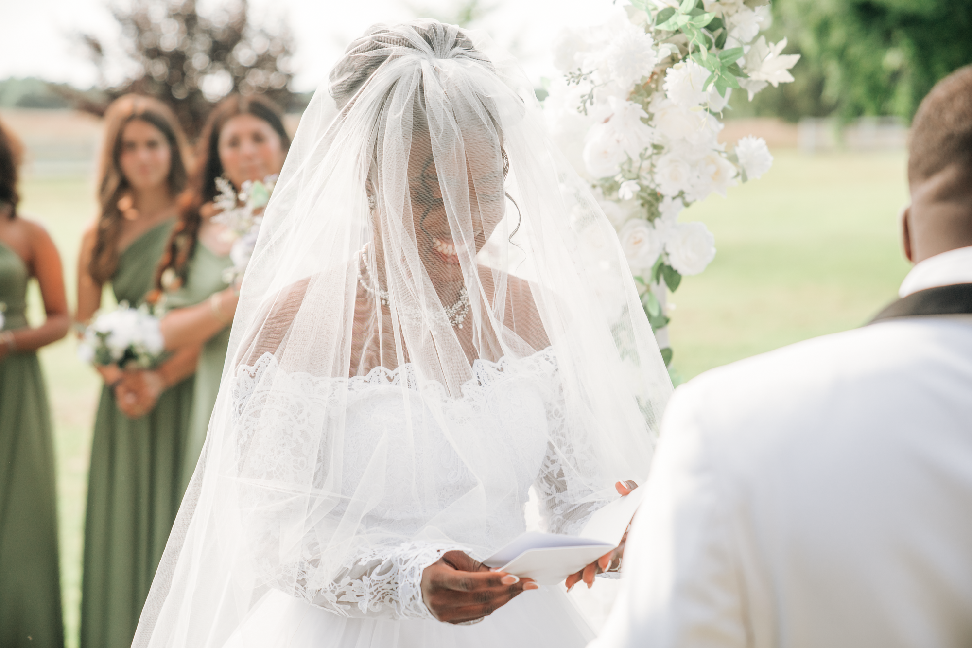 A bride with a lace dress and veil is smiling during her wedding ceremony, reading vows or a speech. Bridesmaids in green dresses are standing behind her holding bouquets.