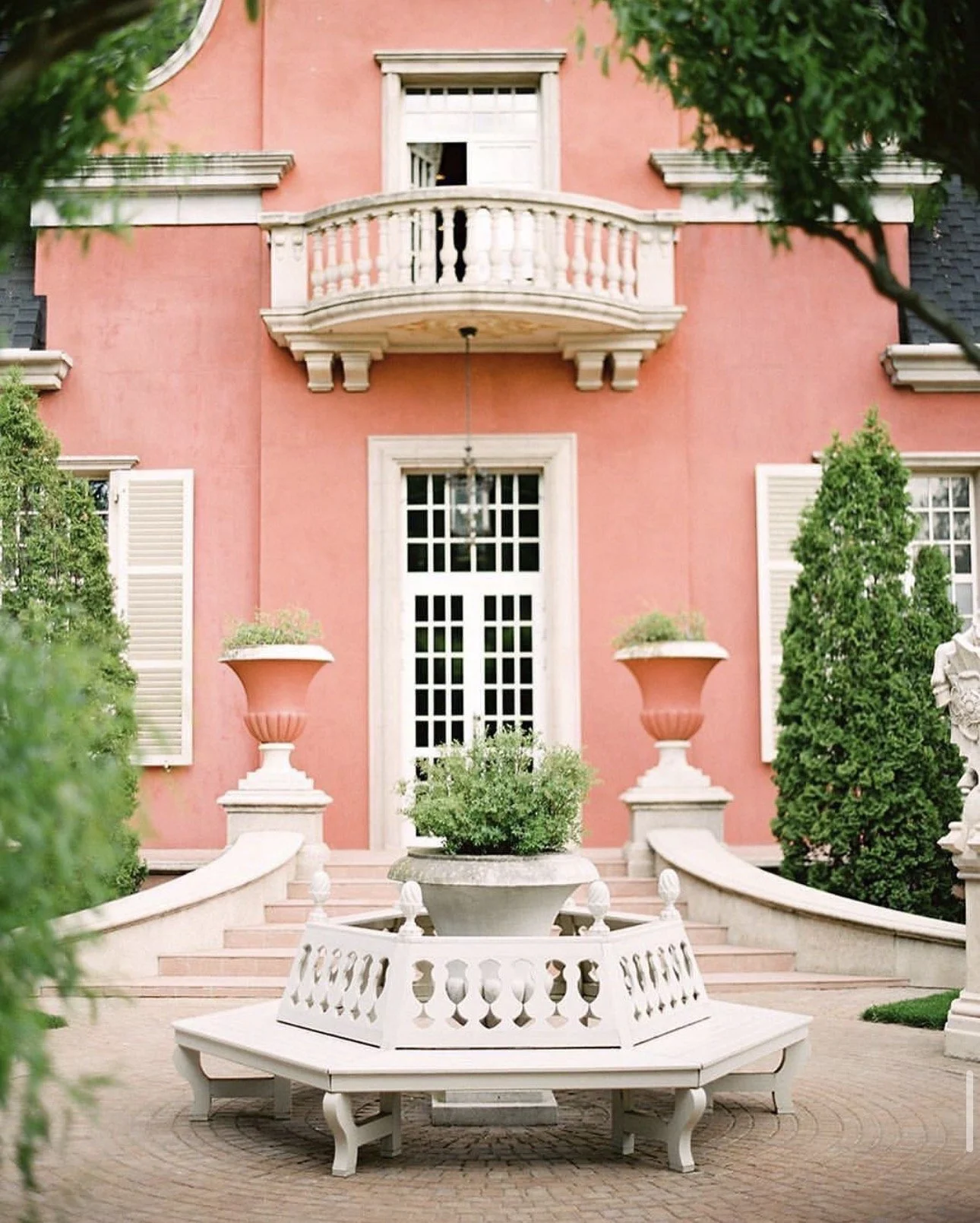 Exterior of a pink house with white trim, a small balcony, and a large white porch with stairs. There are decorative plants in large planters and greenery on the sides.
