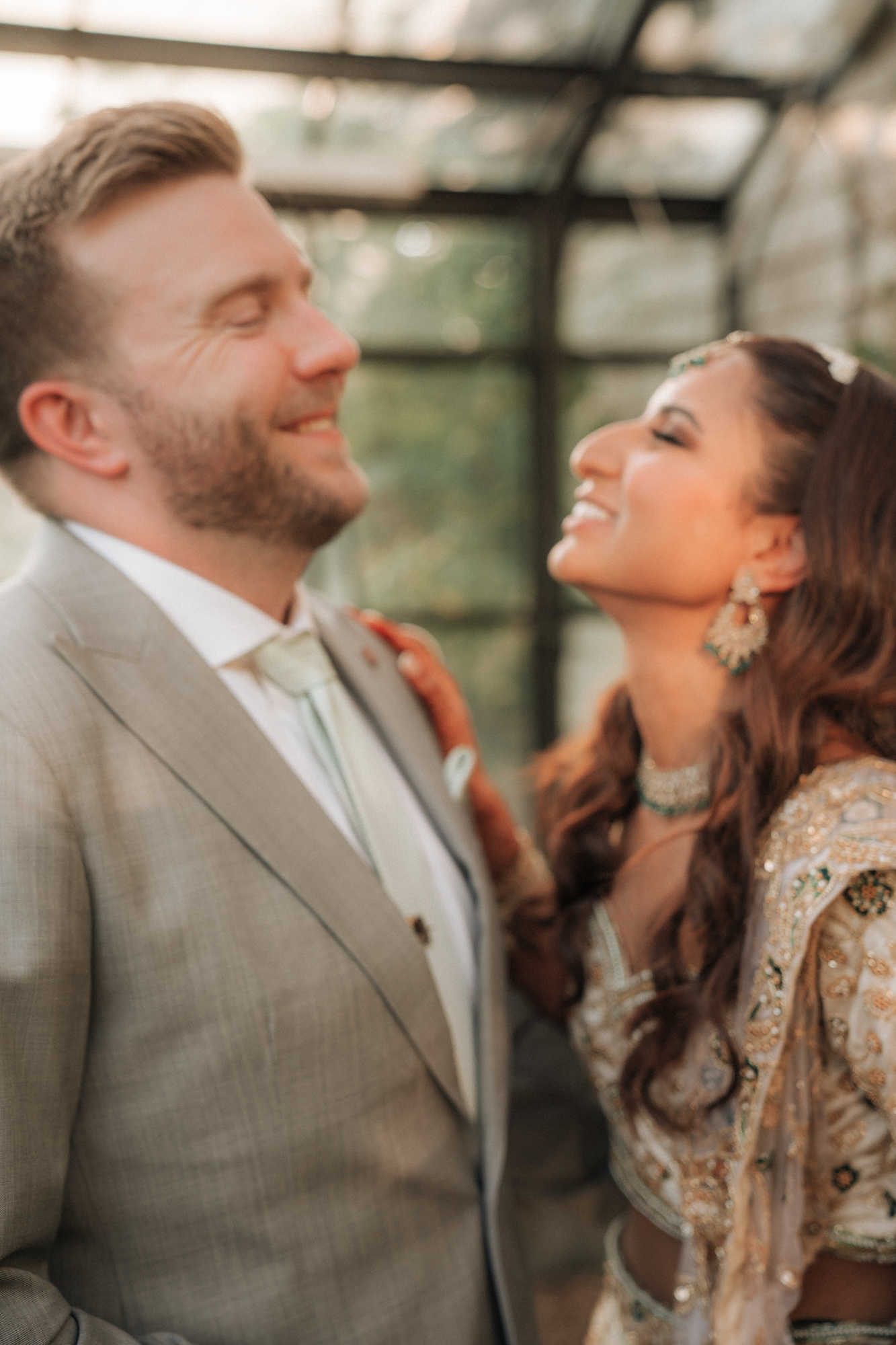 A happy couple, dressed in wedding attire, smiling and looking at each other in an outdoor setting with a glass structure overhead.