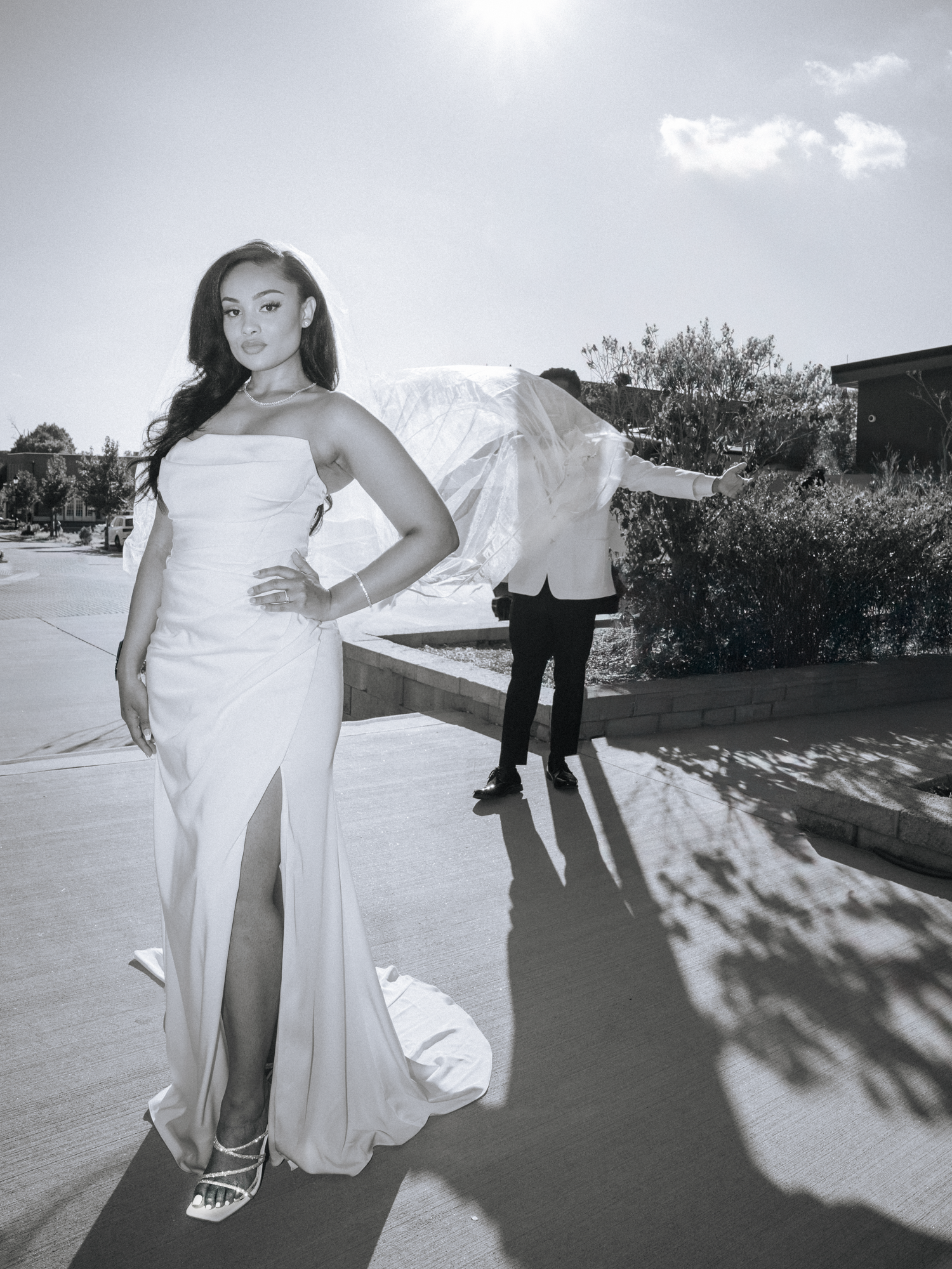 A woman in a white gown standing outdoors with her hand on her hip, with a person in the background adjusting a white suit and umbrella, in black and white photo.