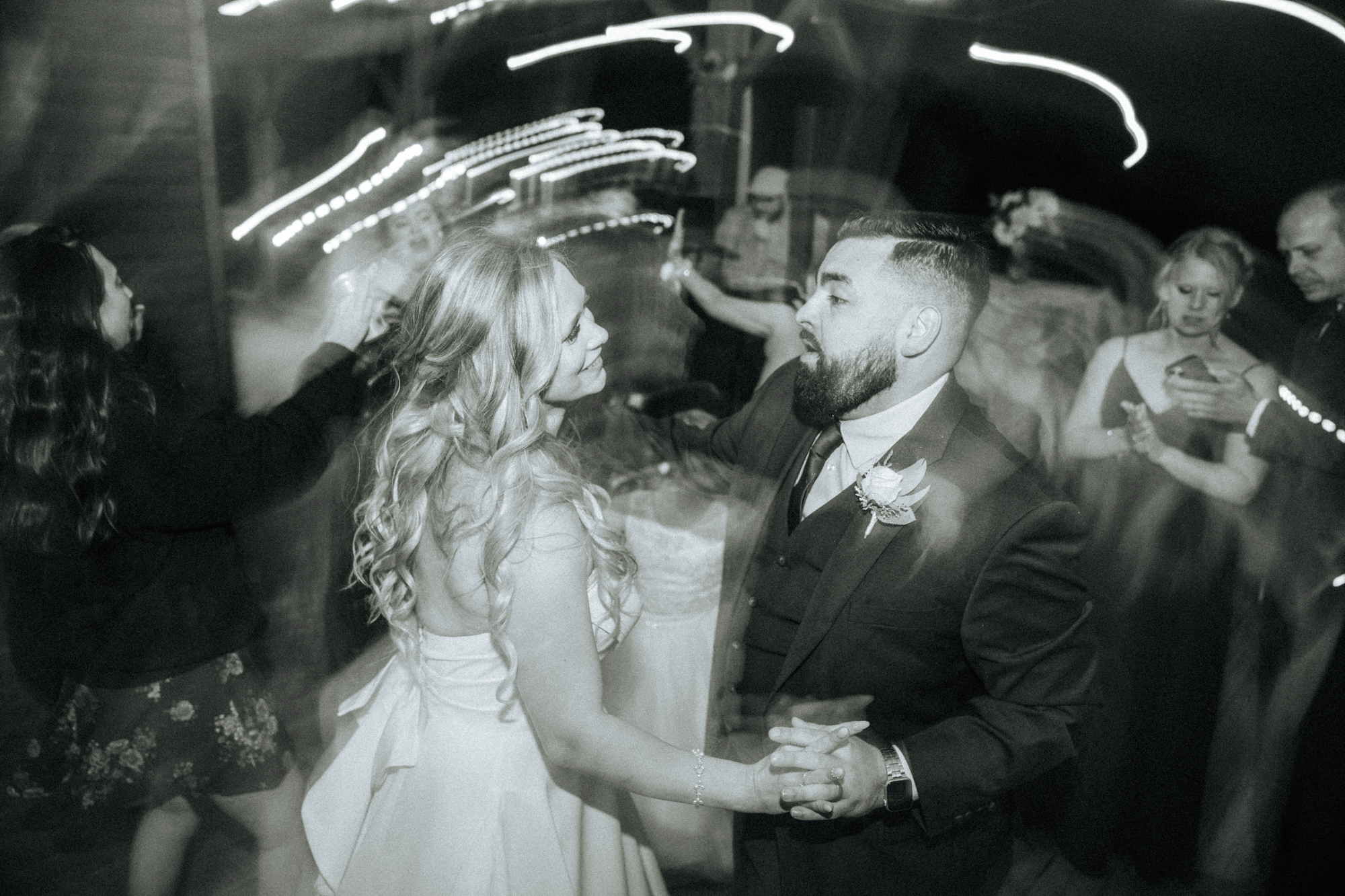 A black and white photo of a wedding reception with a bride and groom dancing closely, surrounded by guests in a dimly lit venue with streaks of light in the background.