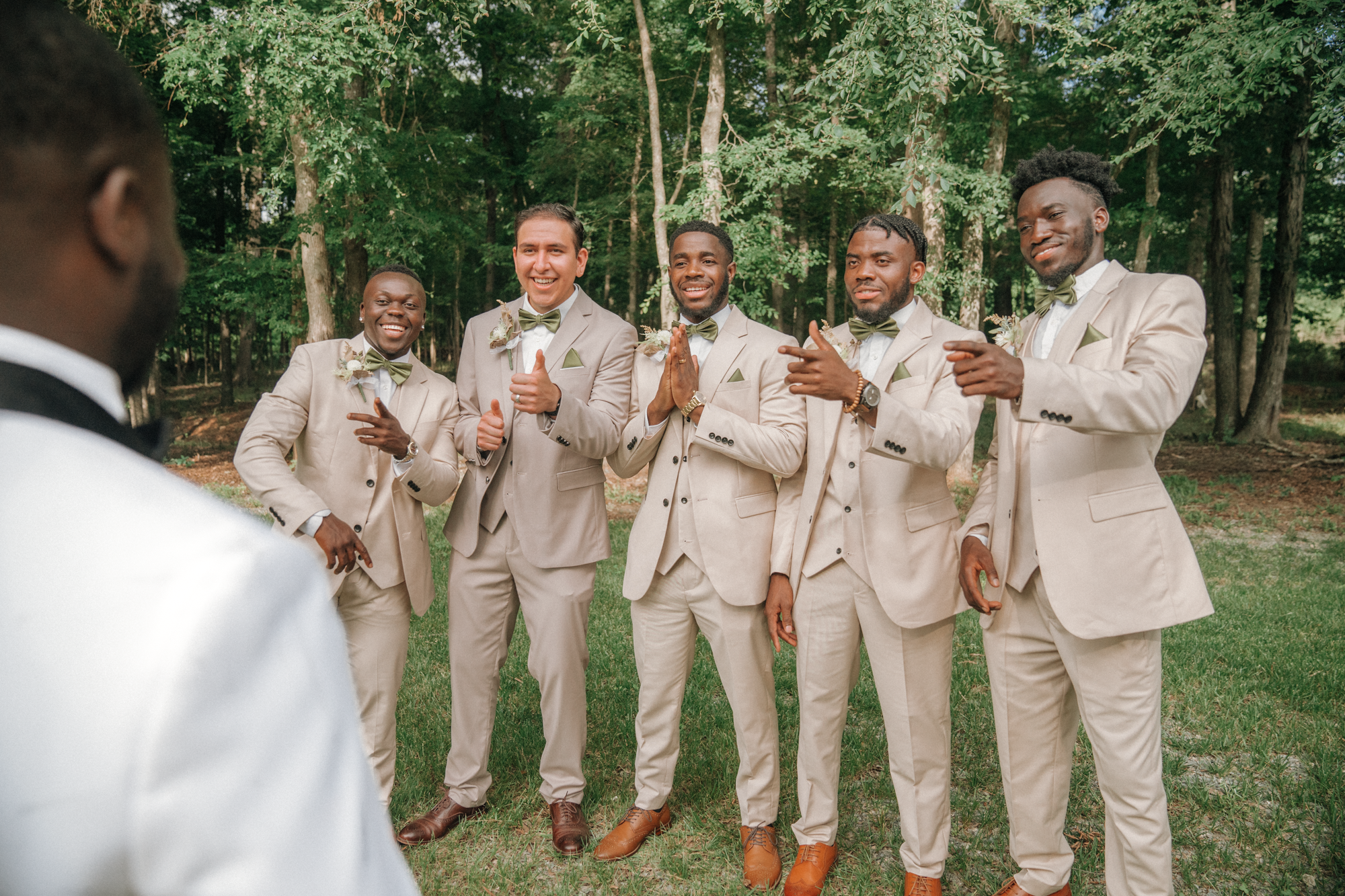 Groom and five groomsmen in beige suits with green bow ties at outdoor wedding, trees in background.