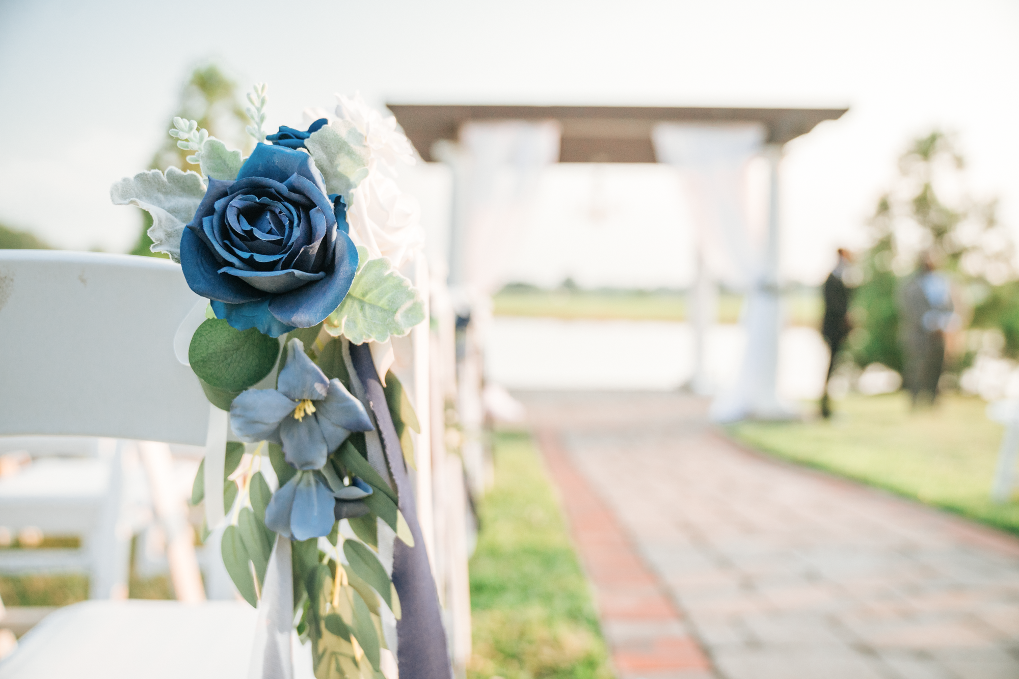 Close-up of a blue and white floral decoration on a white chair at an outdoor wedding altar, with the altar and guests blurred in the background.