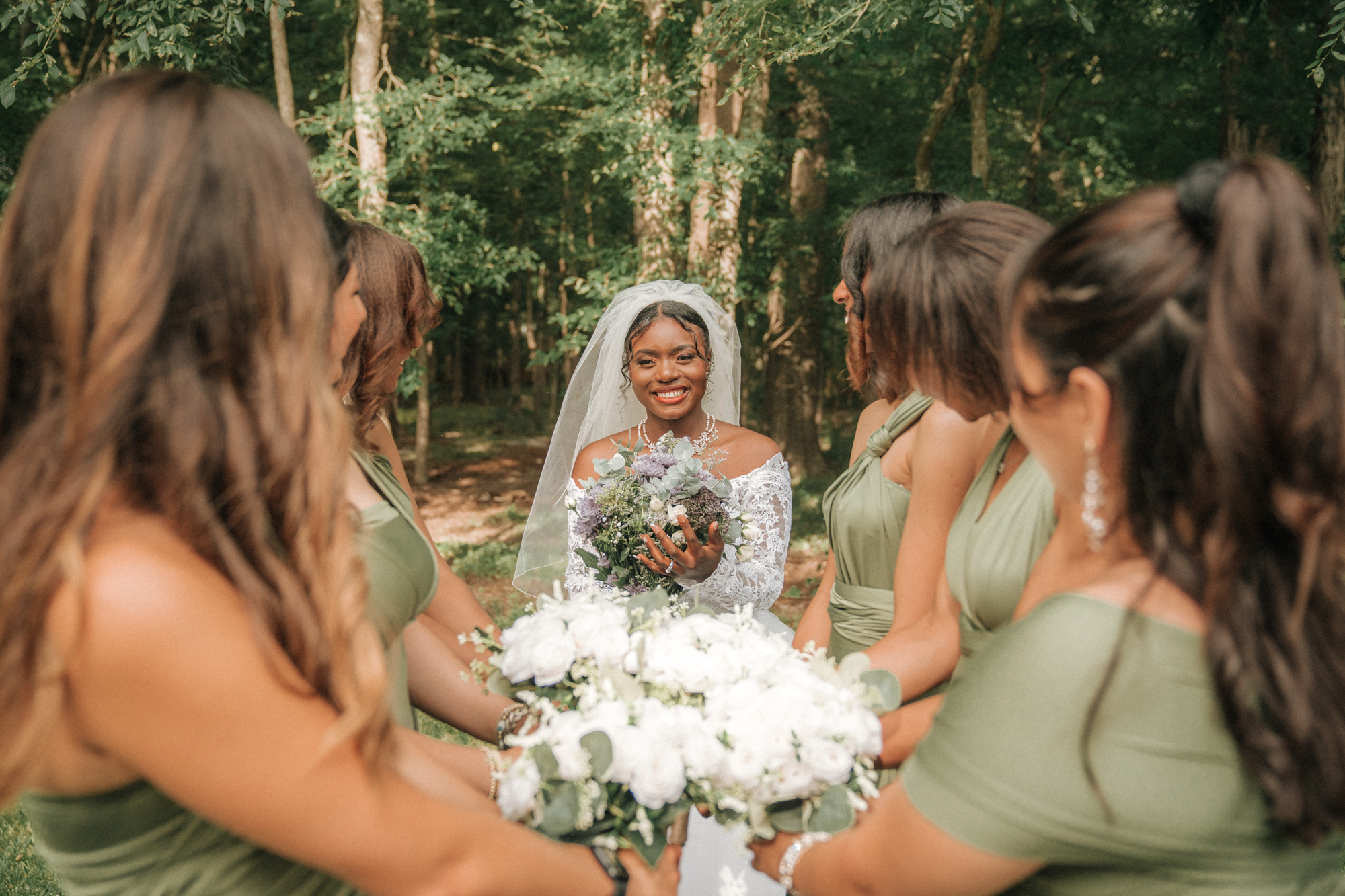 A bride in a lace wedding dress and veil holding a bouquet, smiling at her bridesmaids in green dresses in a wooded outdoor setting.