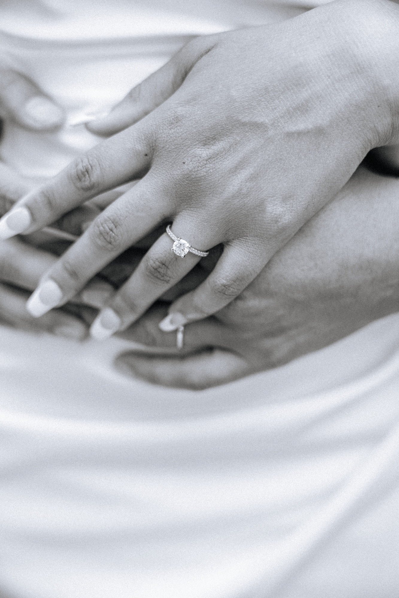 Close-up of a woman's hand with an engagement ring on her finger, resting on a man's hand, both on a white surface.