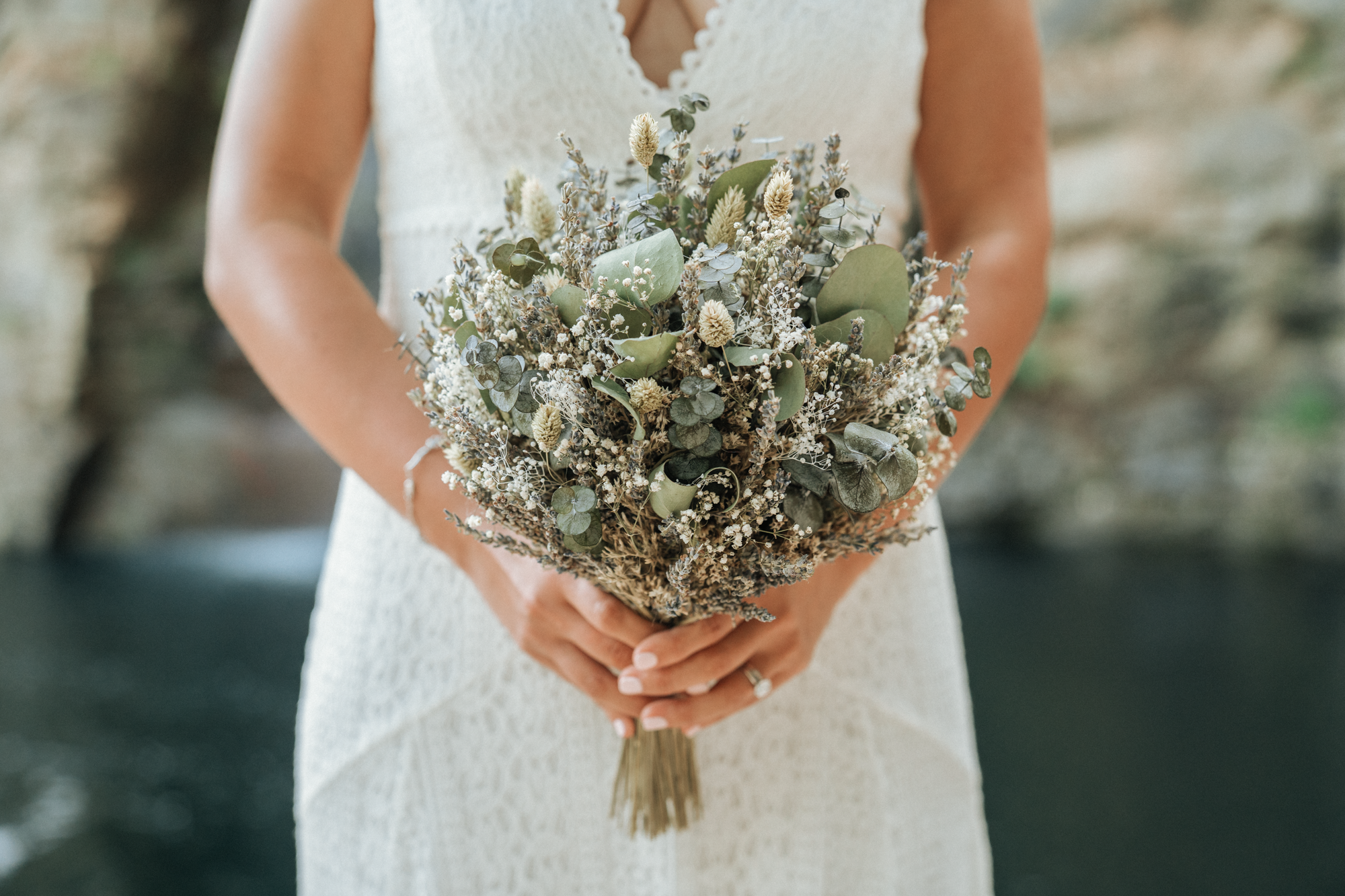 A woman in a white dress holds a bouquet of dried flowers and greenery, standing outdoors with blurred natural background.