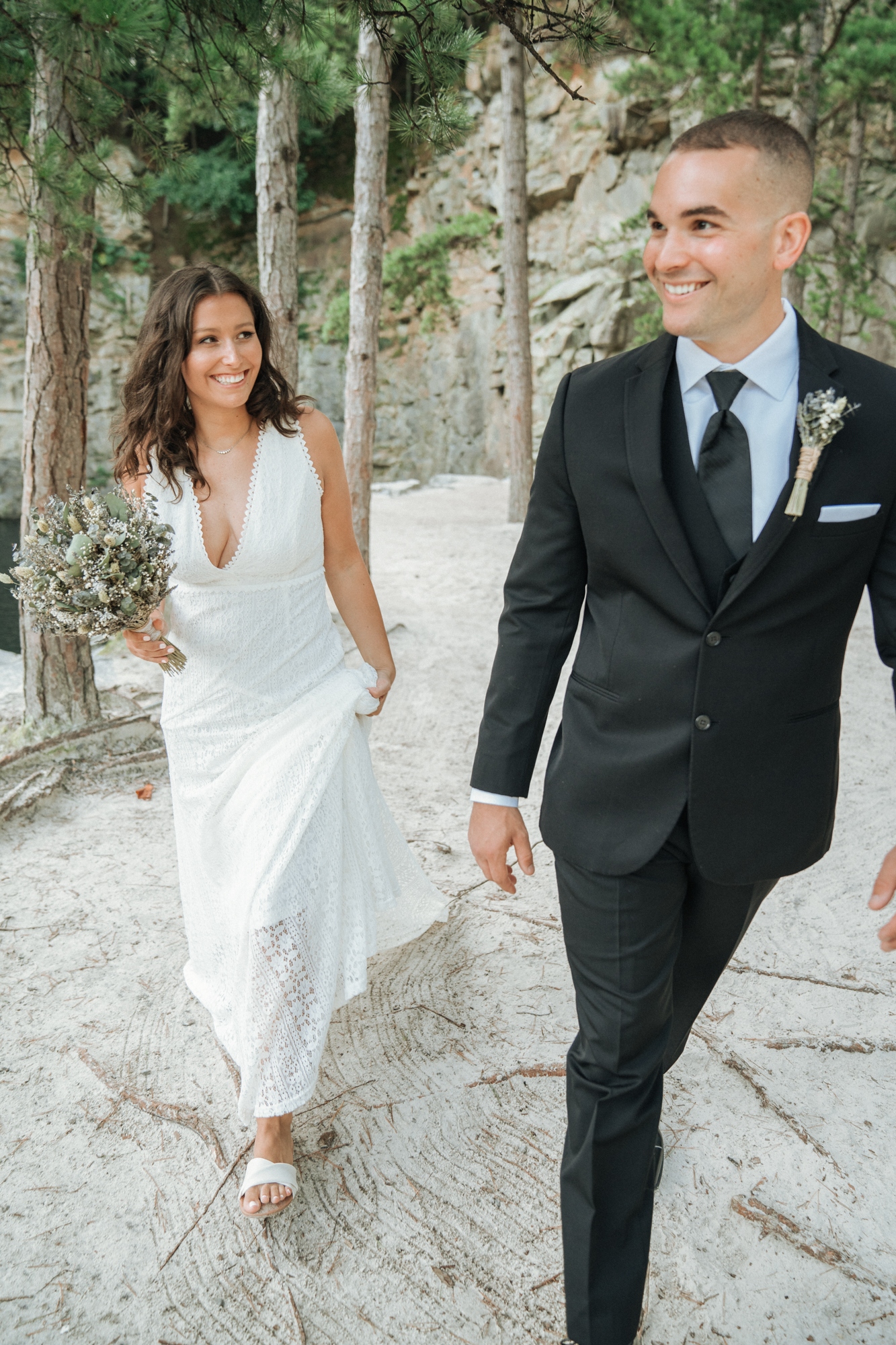 A bride in a white lace dress holding a bouquet and walking on sandy ground next to a groom in a black suit, both smiling in a forested outdoor setting.