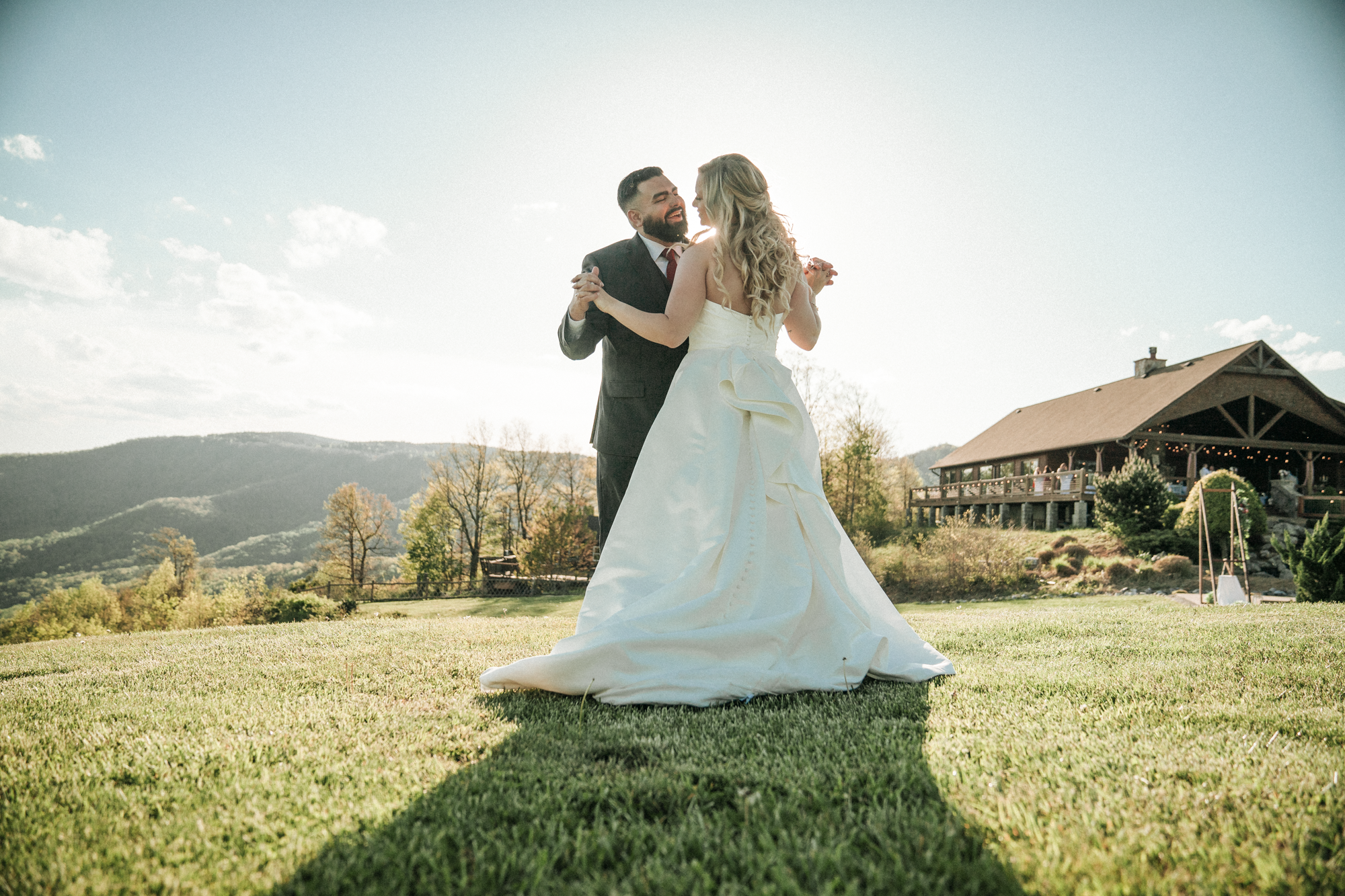A newlywed couple dancing outdoors on a grassy field with scenic mountains in the background and a rustic building on the right.