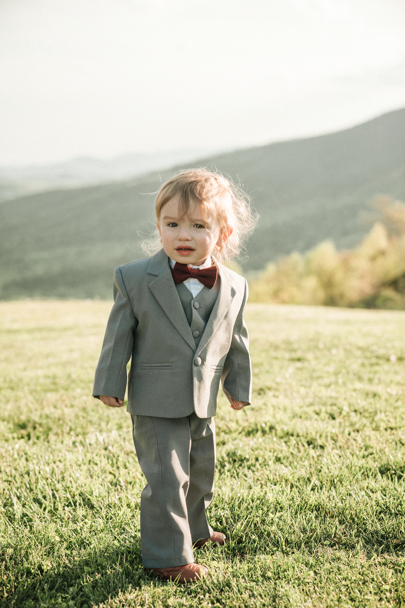 A young child in a gray suit with a red bow tie standing outdoors on grass with a mountainous landscape in the background.