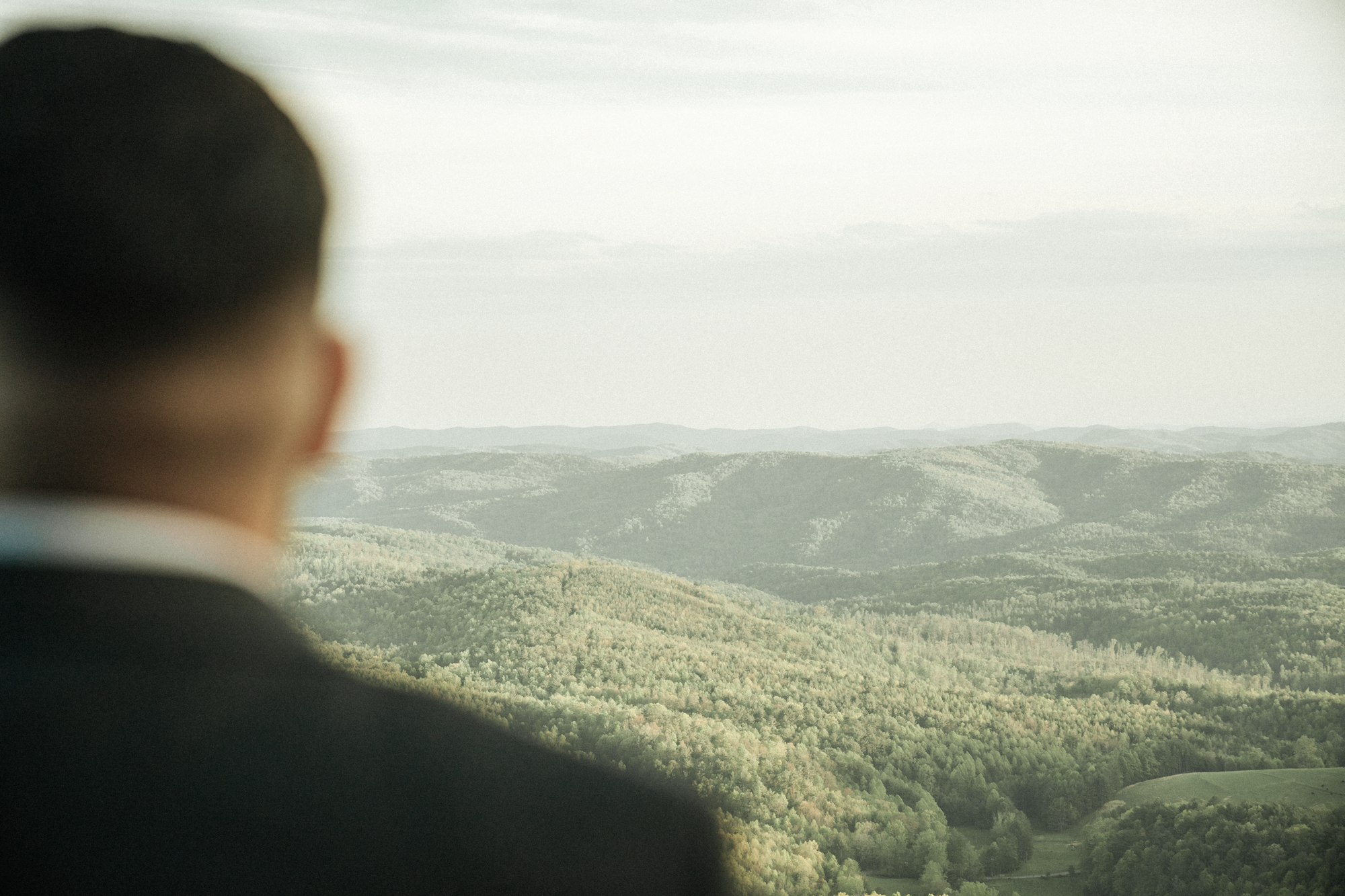 Person in a suit looking at a vast view of layered mountains and green hills from a high vantage point.