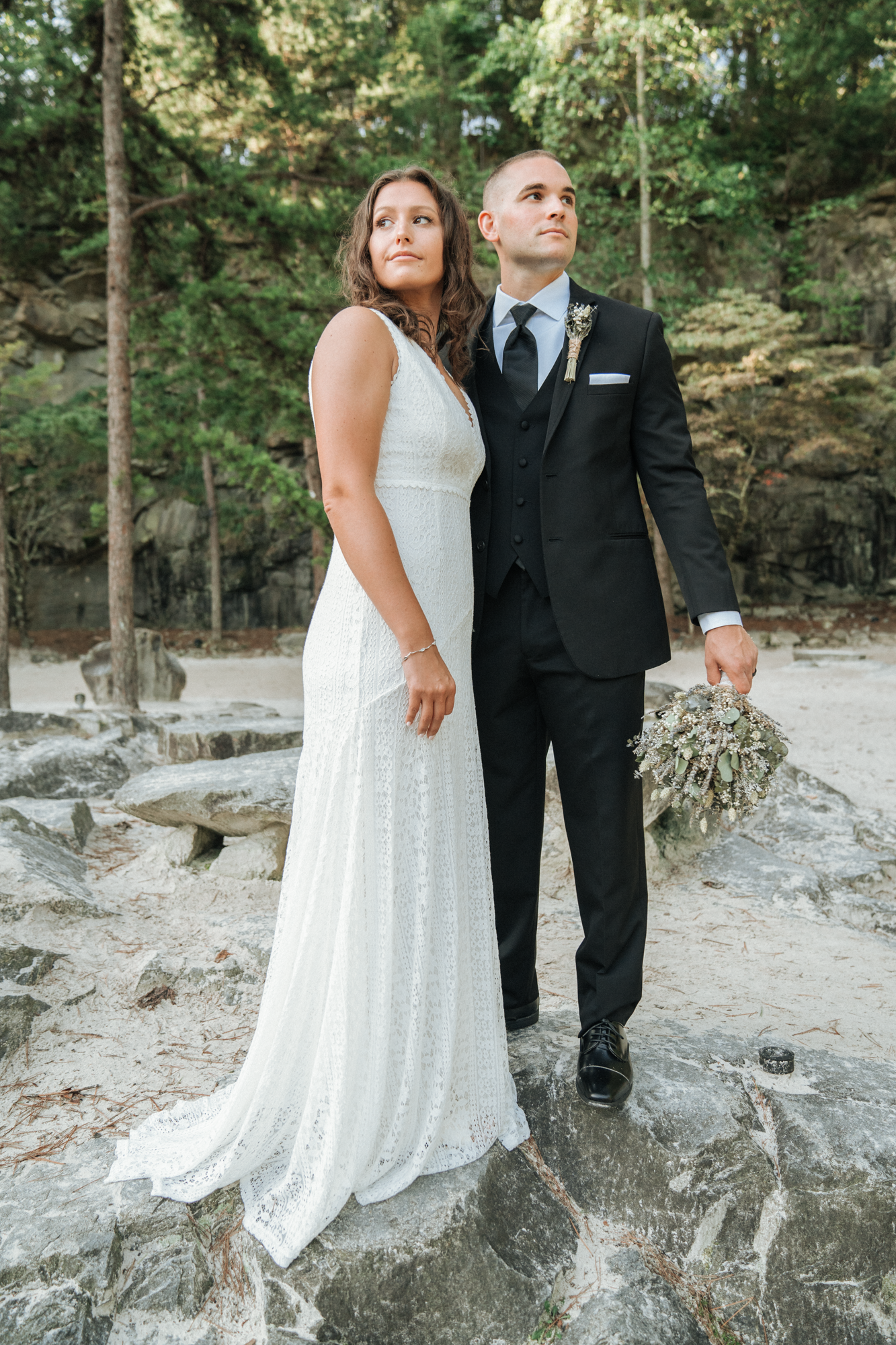 A bride and groom standing outdoors on rocks with trees in the background, dressed in wedding attire, holding a bouquet.