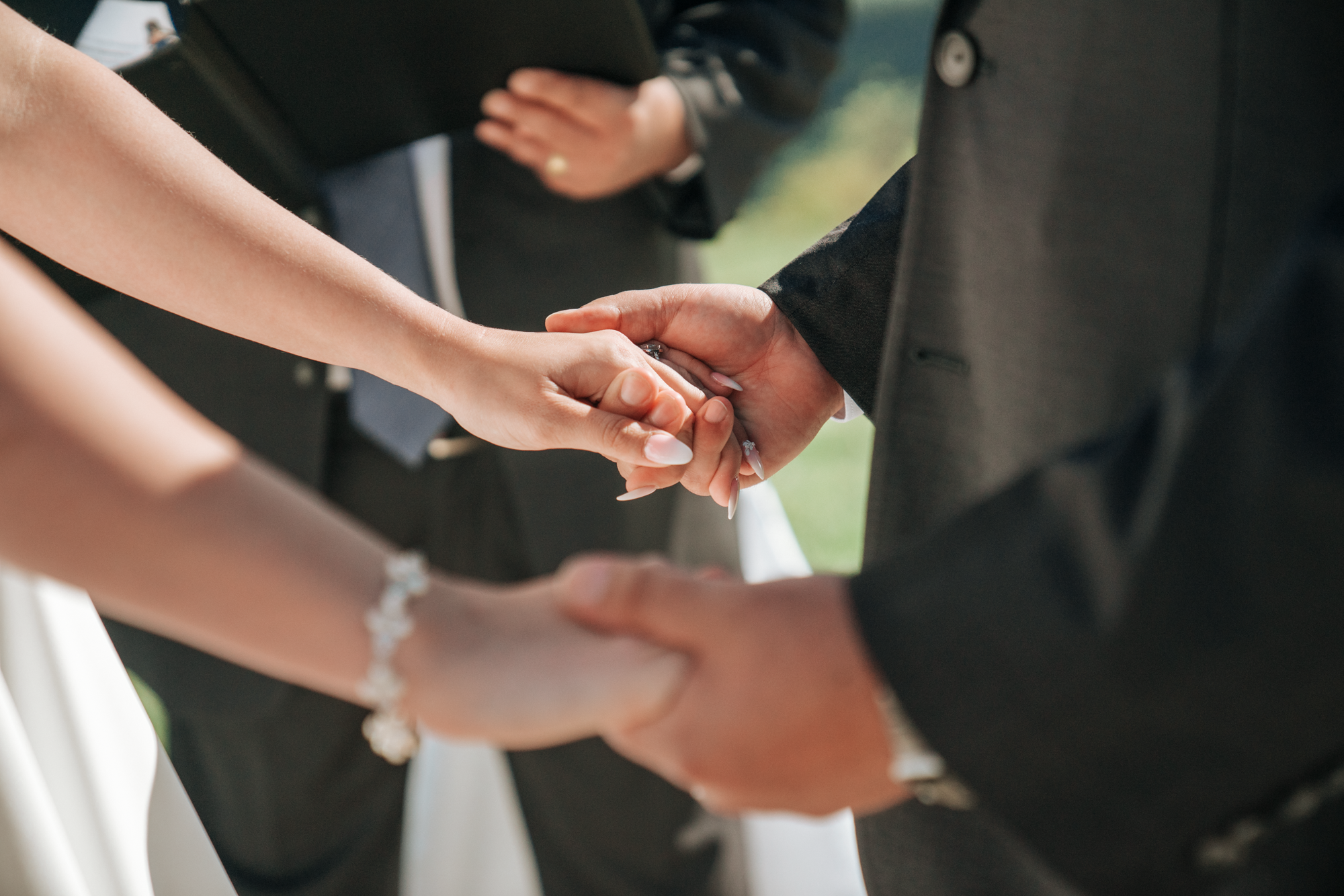 Close-up of a bride and groom holding hands during a wedding ceremony, with an officiant in the background.