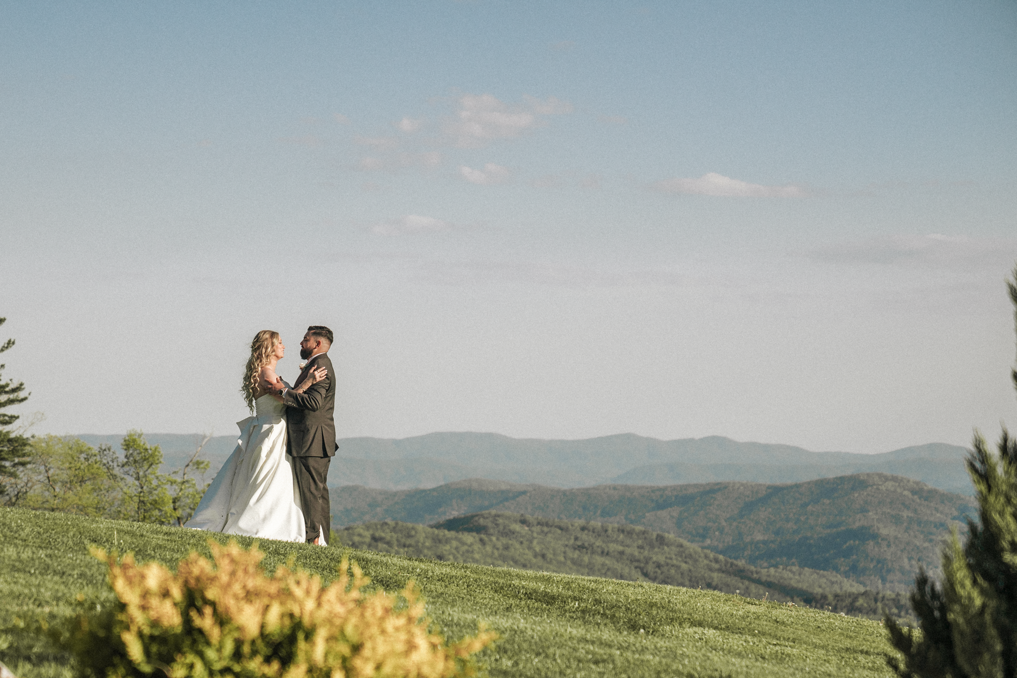 A bride and groom stand facing each other on a grassy hill with a mountain landscape in the background during a wedding photoshoot.
