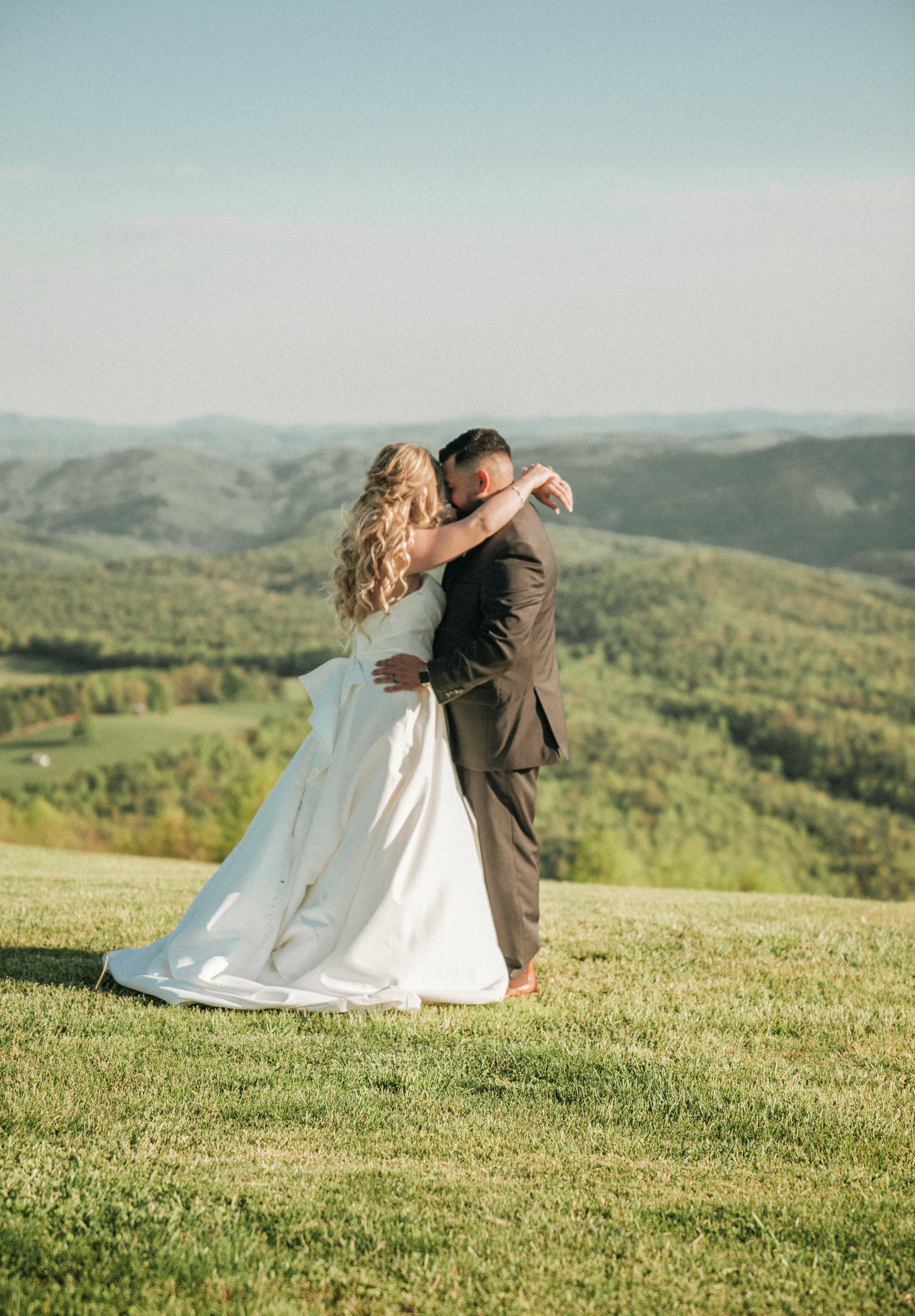 A bride and groom embracing outdoors on a grassy hill with rolling green hills and a clear blue sky in the background.