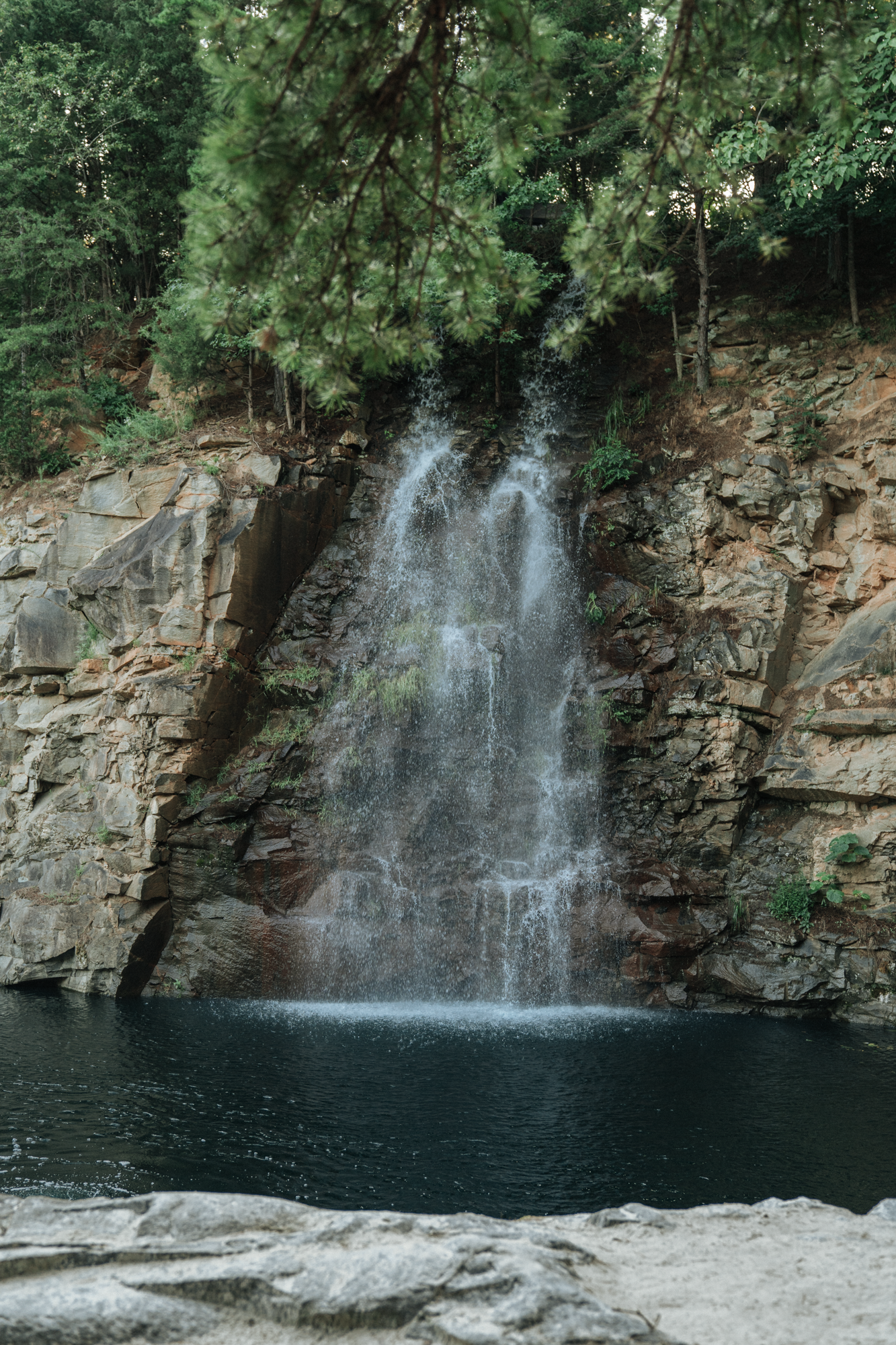 A waterfall flowing down a rocky cliff into a dark pool of water, surrounded by green trees.
