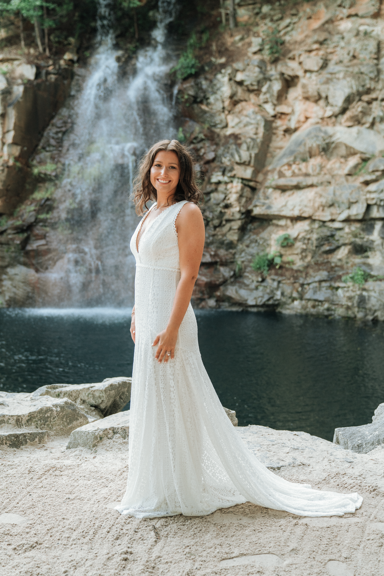 A smiling woman in a white lace dress standing on rocks near a waterfall and pond.