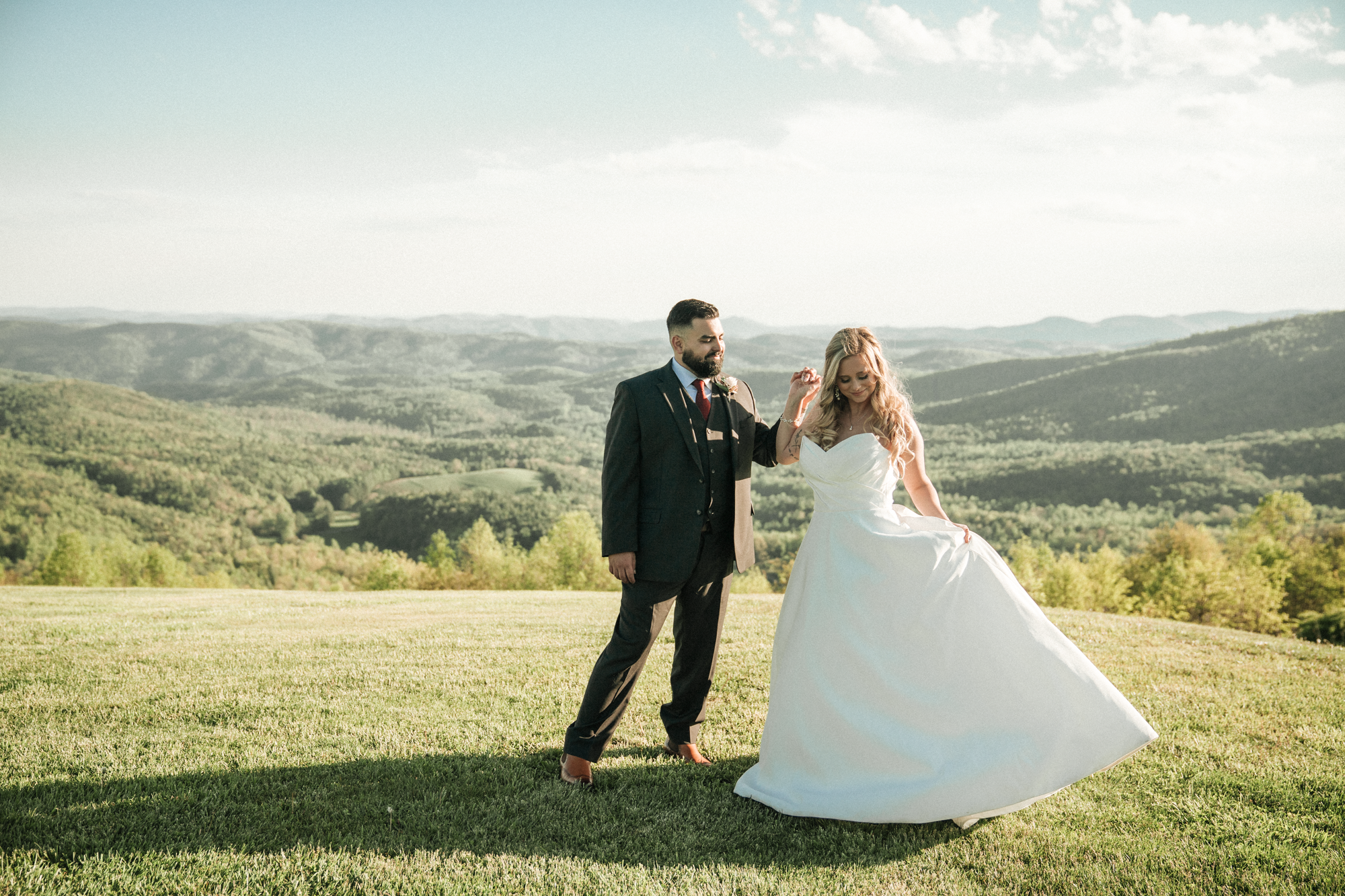 A couple dancing outdoors on a grassy field with rolling hills and mountains in the background, the woman in a white wedding dress and the man in a dark suit.