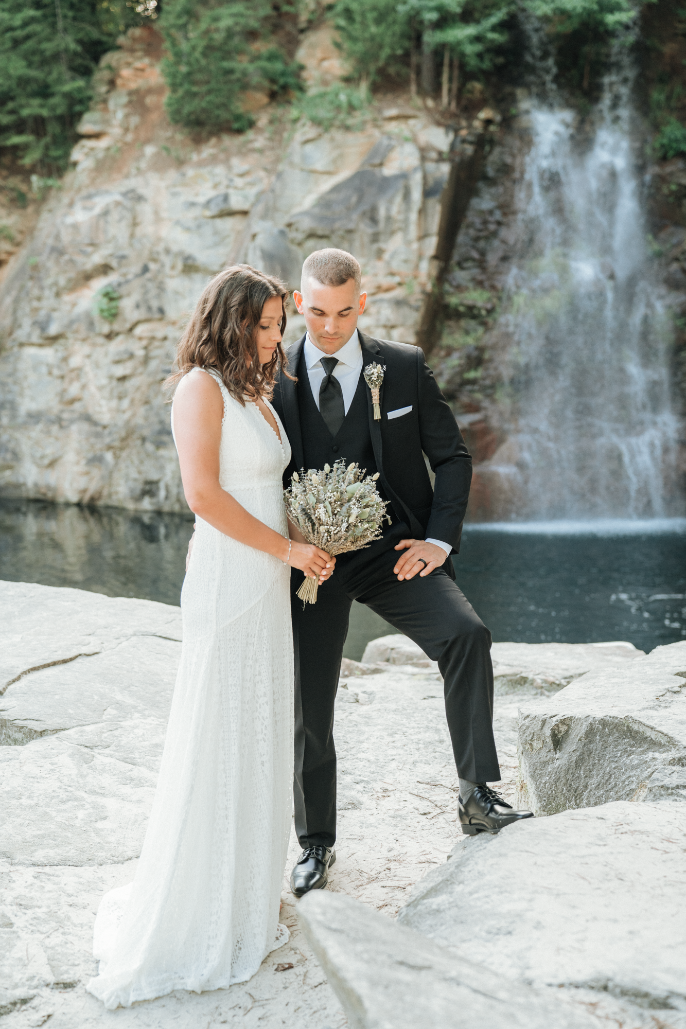 A bride and groom stand together on rocks near a waterfall, with the bride holding a bouquet and the groom dressed in a black suit, in an outdoor scenic setting.
